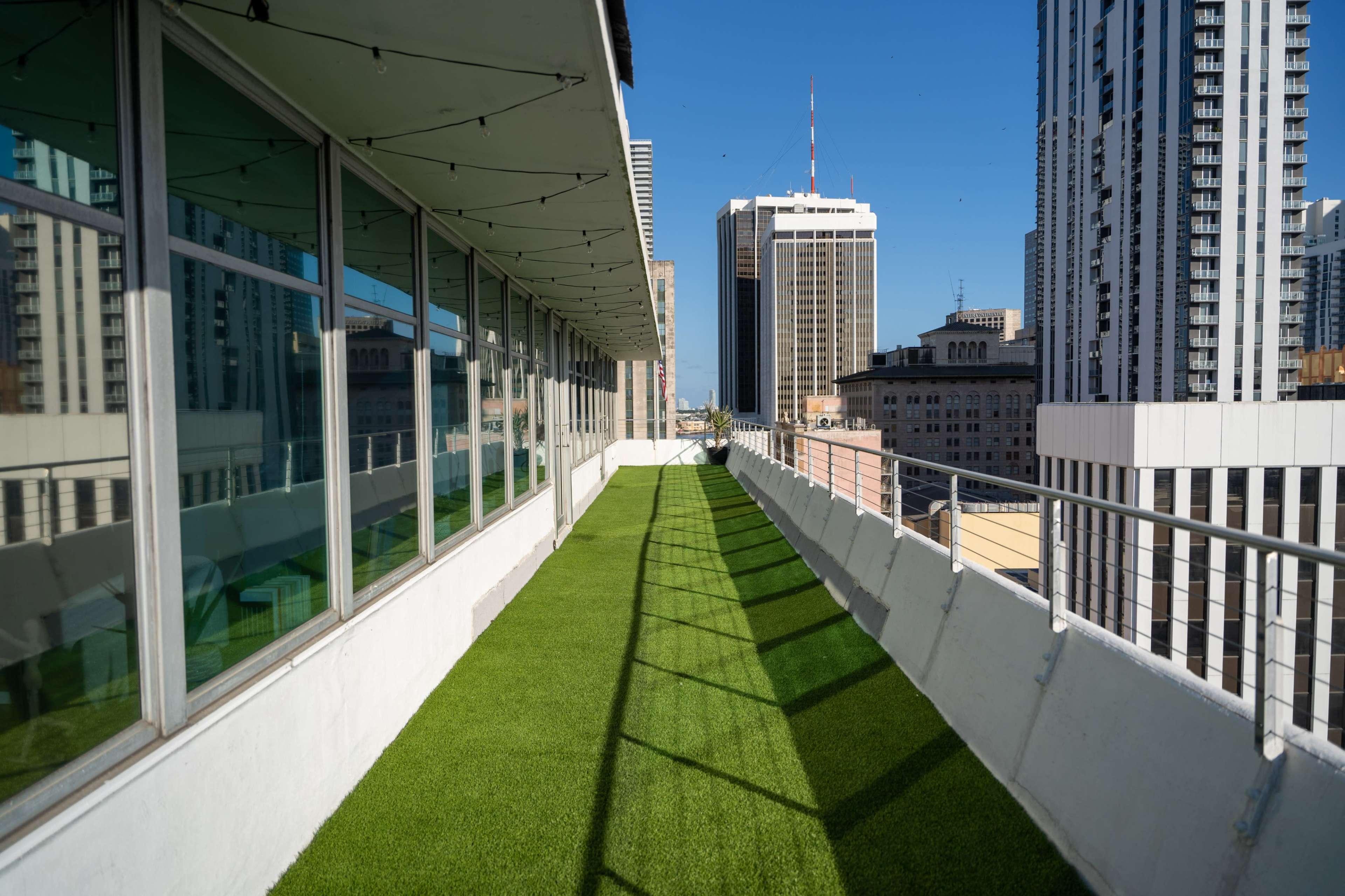 A narrow balcony with artificial grass extends along a building, offering views of surrounding skyscrapers against a clear blue sky.