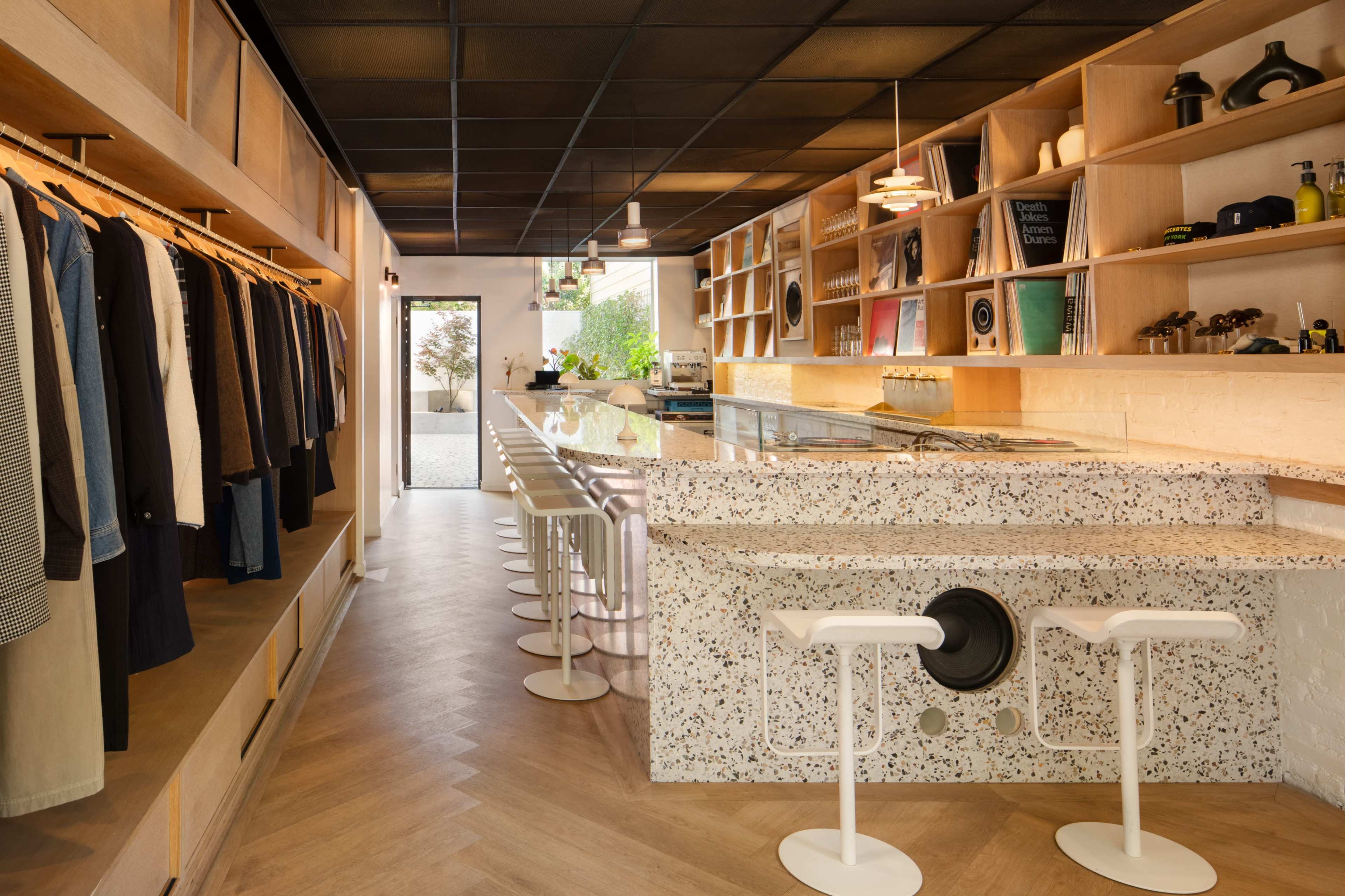 The interior of a stylish boutique features a long bar with high stools, clothing racks on the side, and a modern design with light wood and speckled counter surfaces.