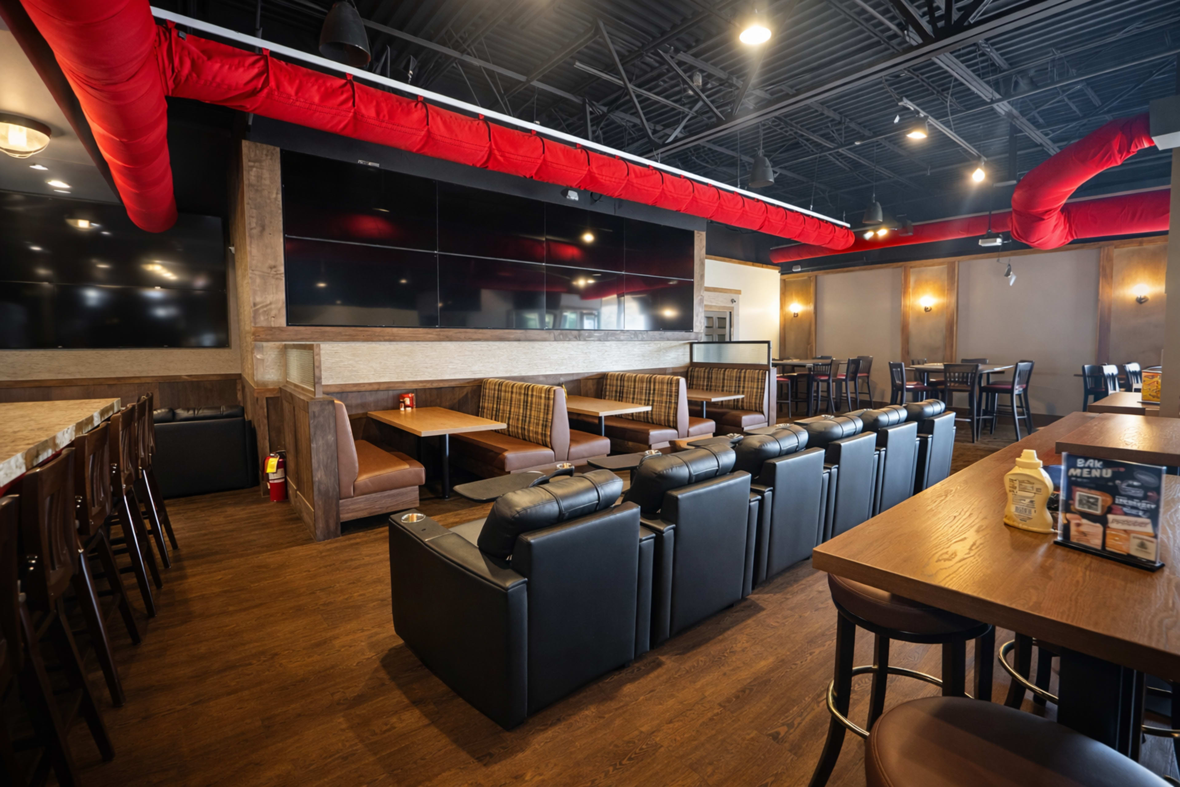 The image shows a modern restaurant interior featuring a mix of booth and table seating, large wall-mounted screens, and exposed red ductwork.