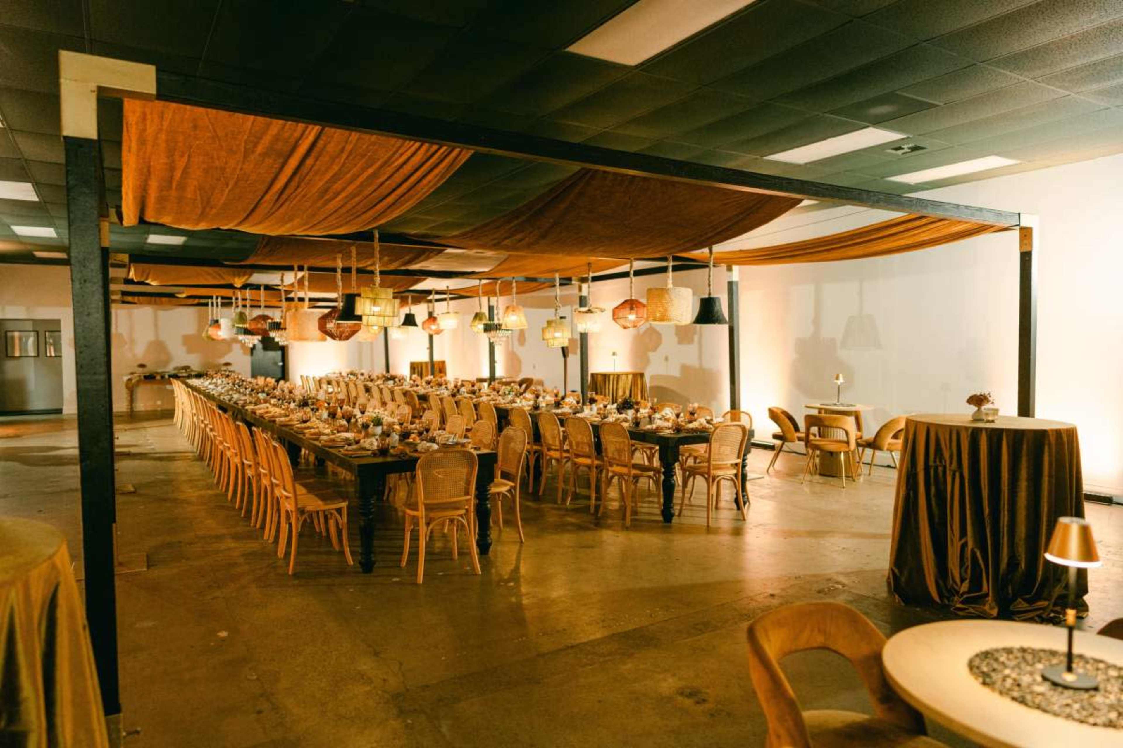 A large banquet hall features long tables set for an event, with decorative drapes and pendant lighting hanging from the ceiling.