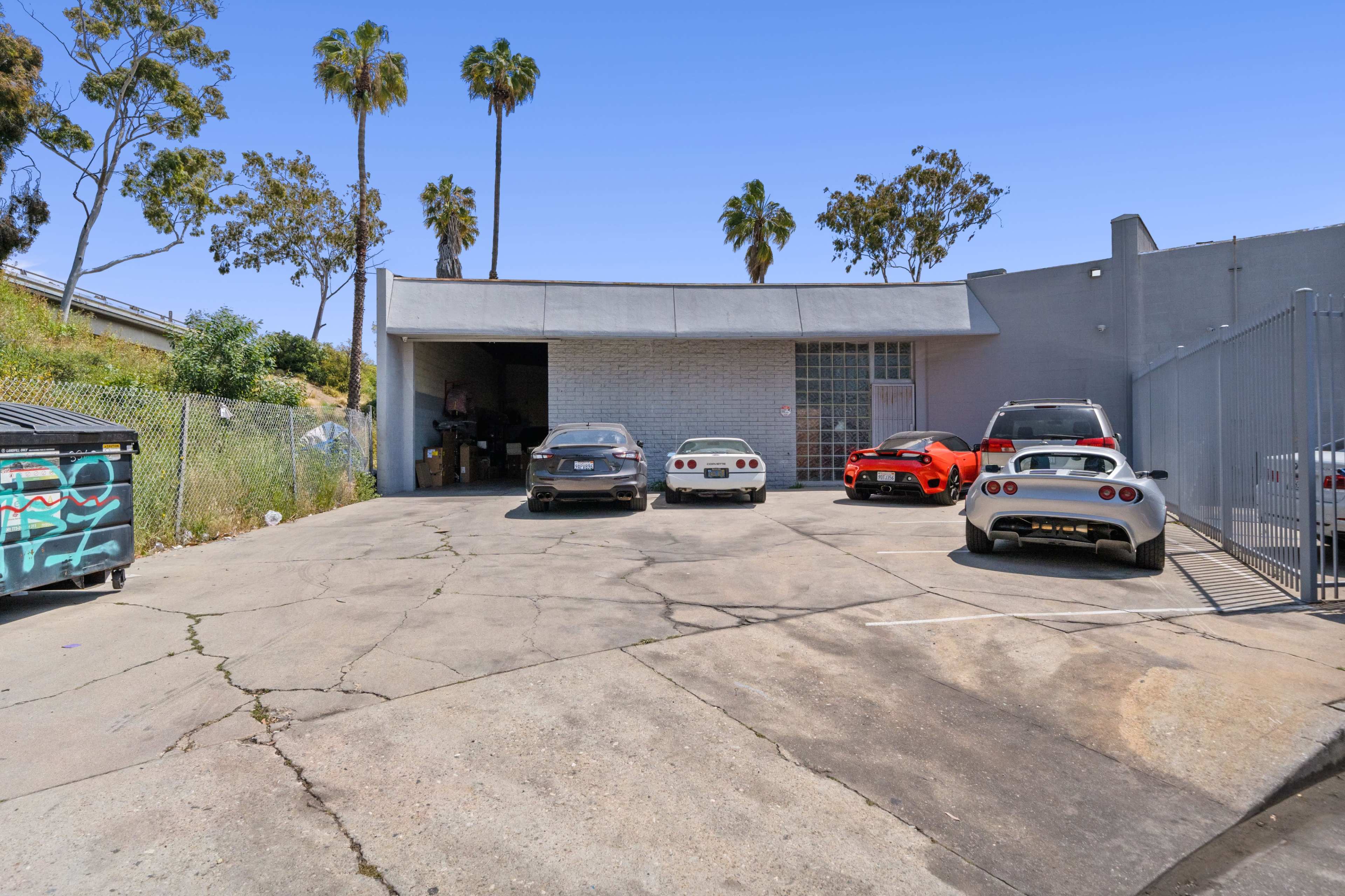 A parking lot behind a building features several cars, including a red sports car and a white vehicle, surrounded by palm trees.