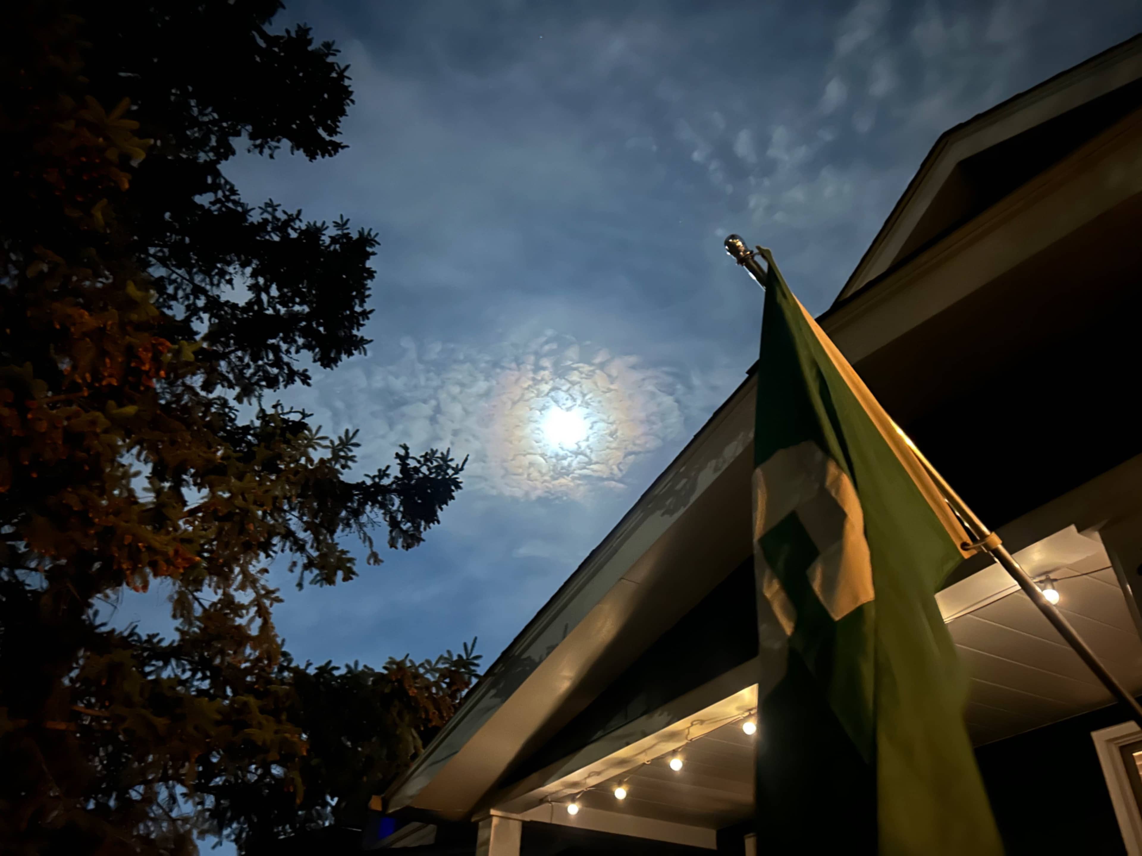 A green flag waves in front of a house under a partly cloudy night sky illuminated by the moon.