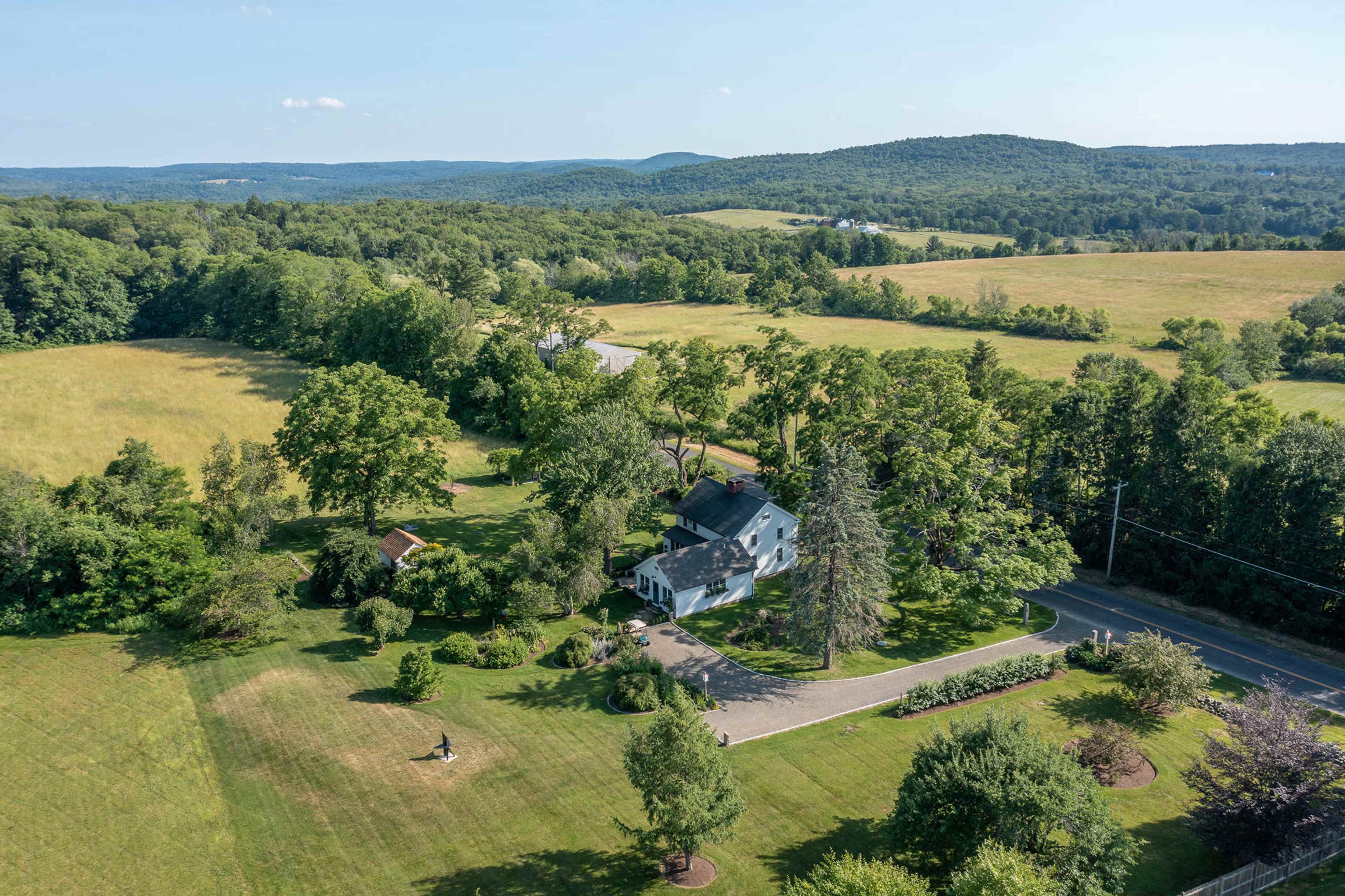 An aerial view shows a house surrounded by lush trees and fields, with a winding road visible nearby.