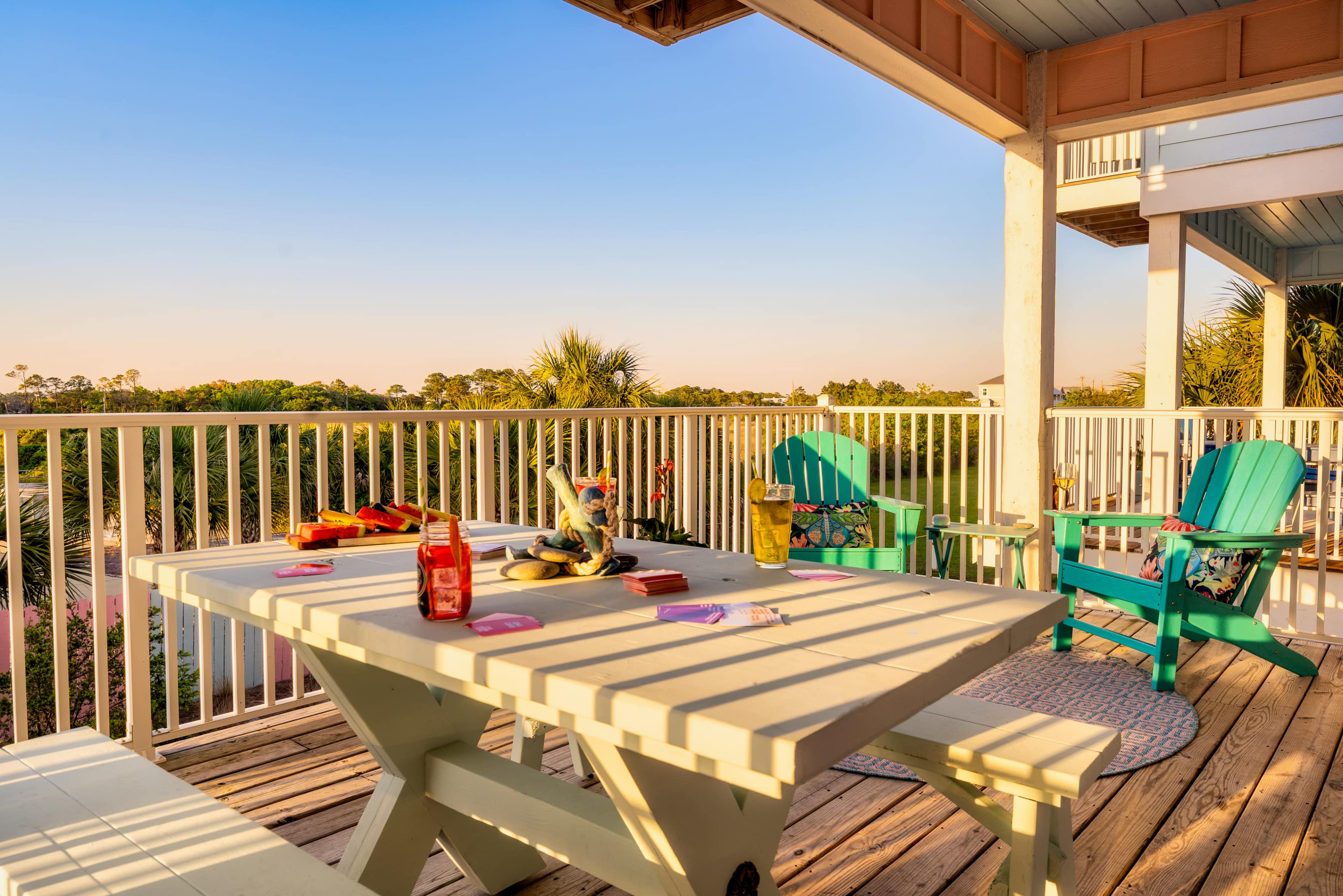 A wooden deck features a table set with condiments and snacks, surrounded by teal chairs and a view of palm trees under a clear sky.