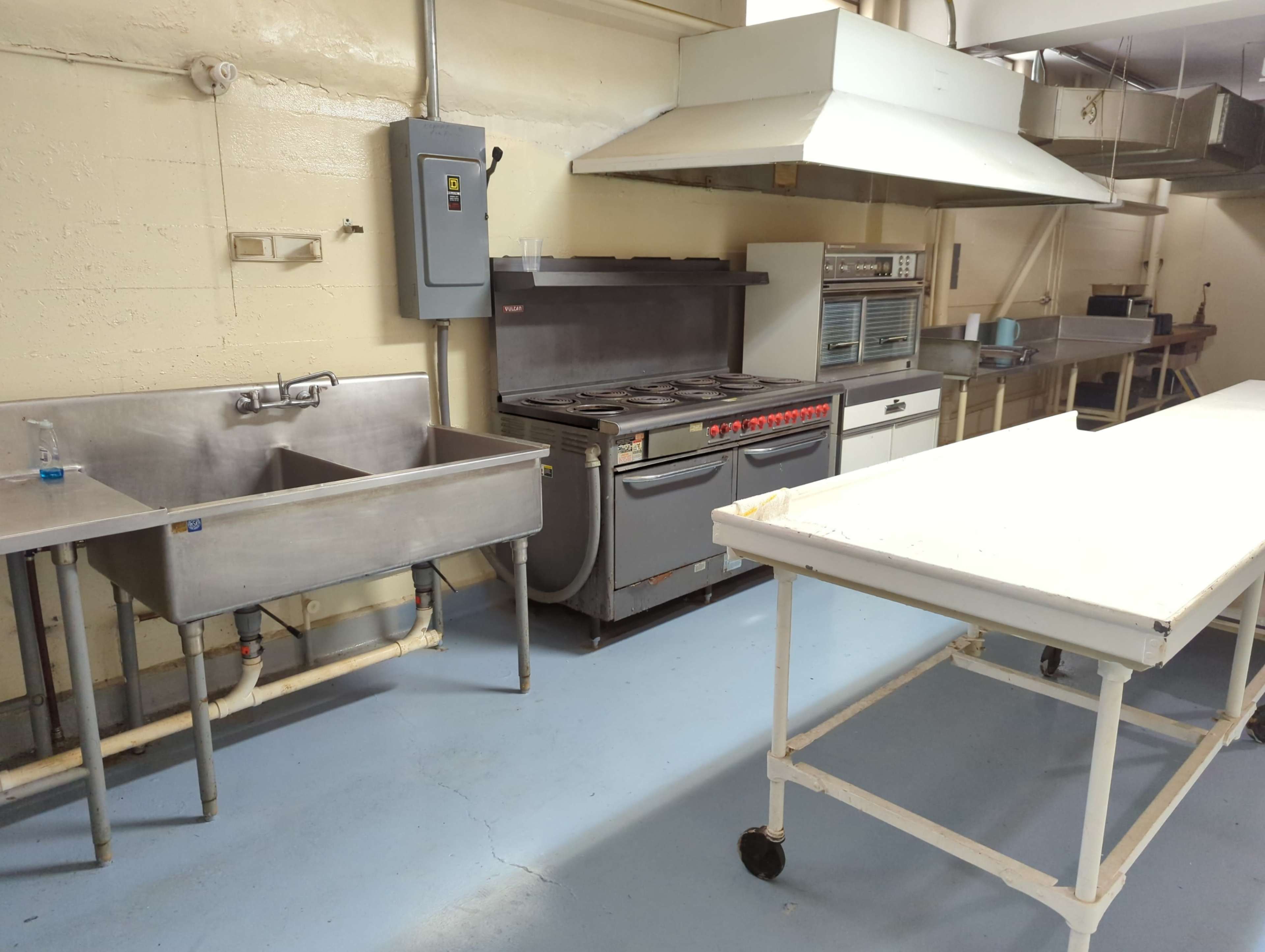 A commercial kitchen equipped with a stainless steel sink, a gas range with multiple burners, a metal work table, and ventilation hoods.