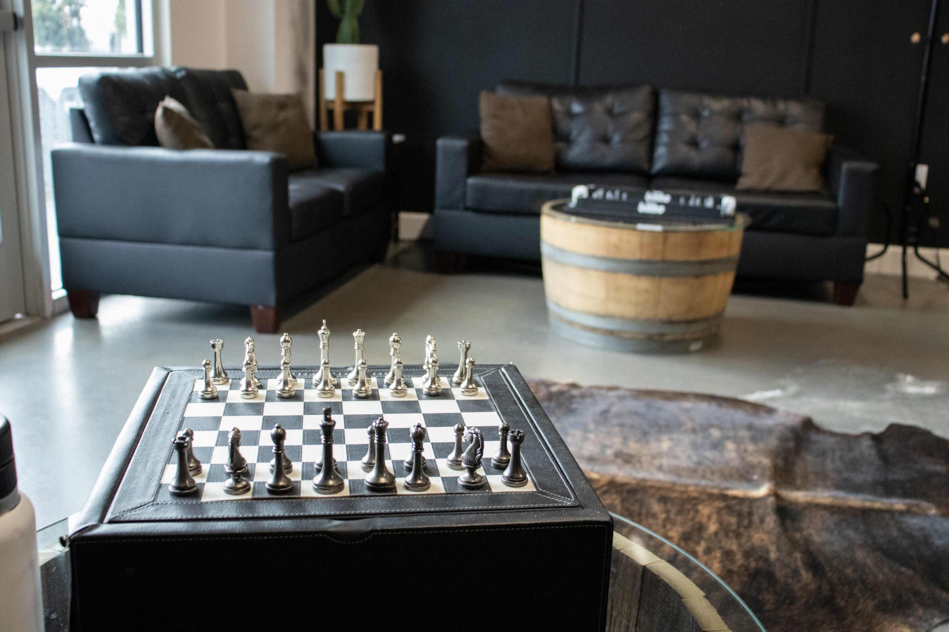 A chess set is positioned on a glass table in the foreground of a modern living room featuring a dark sofa and a wooden barrel table.