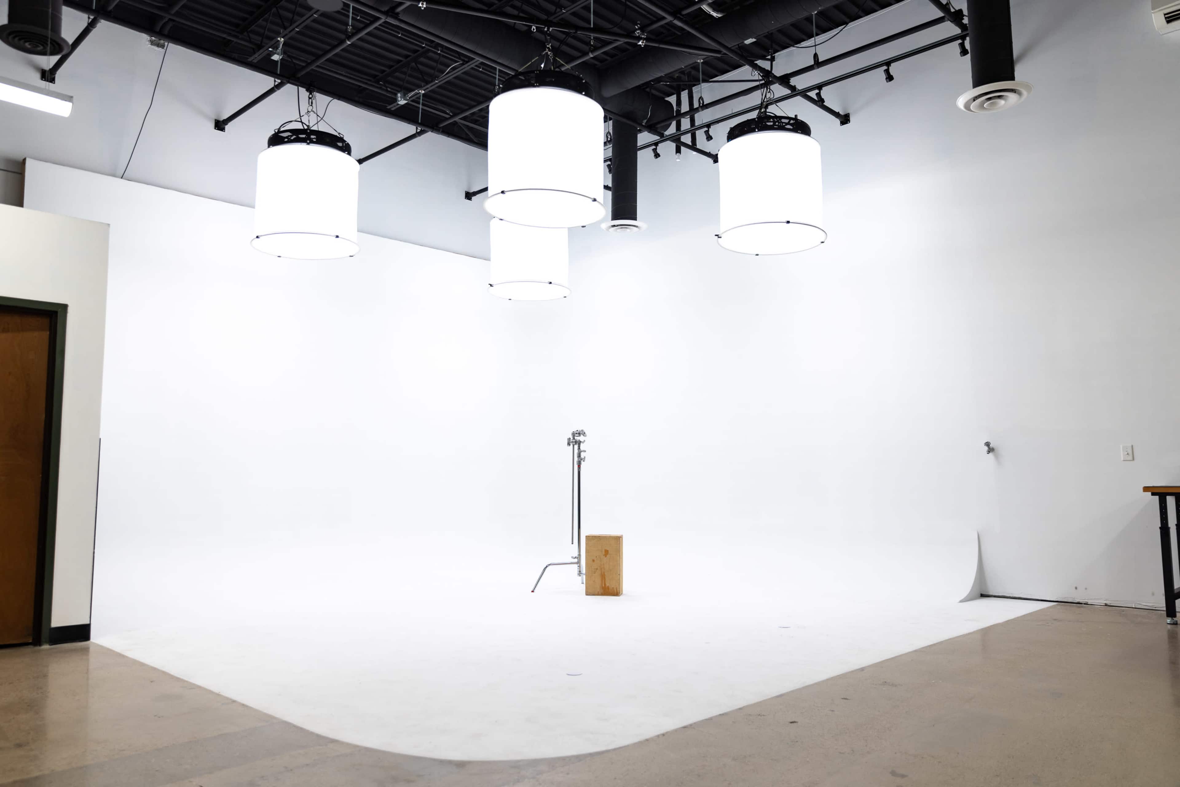 The image shows a spacious, well-lit studio with several large pendant lights hanging from the ceiling, a simple wooden stool on a white backdrop, and a smooth floor.