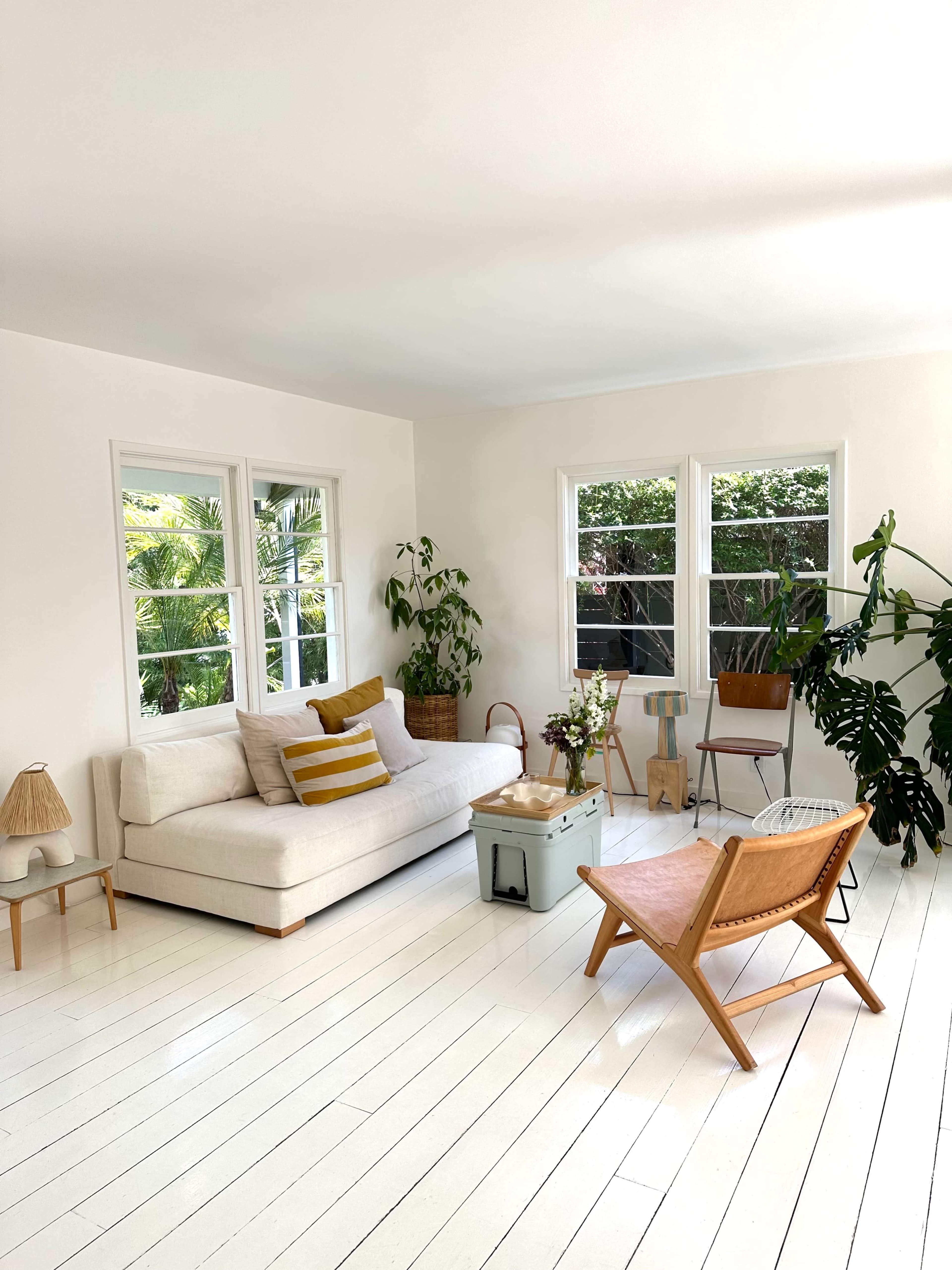 A bright living room features a white sofa, wooden furniture, potted plants, and large windows allowing natural light.