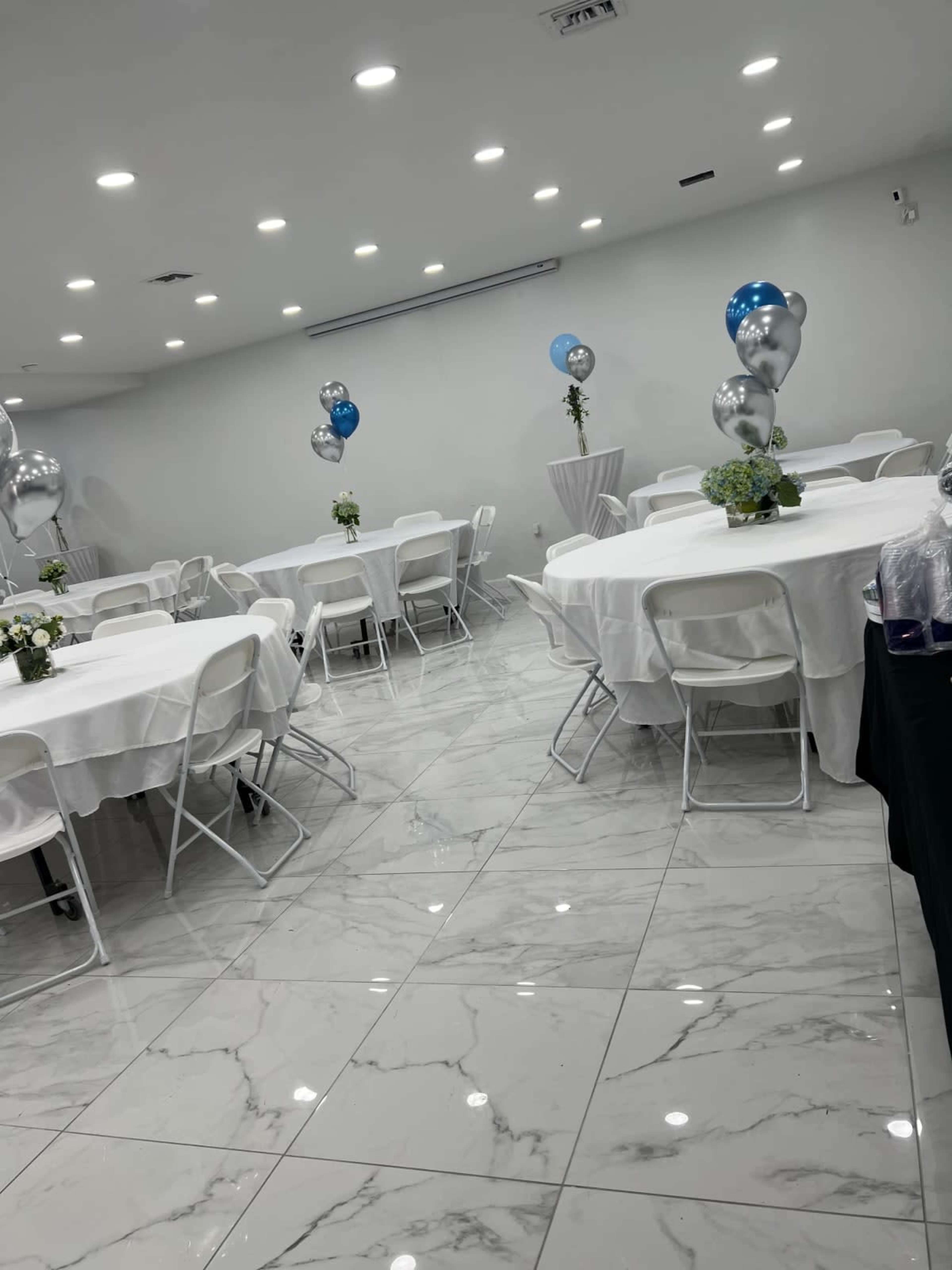 The image shows a banquet hall with white tables covered in tablecloths, surrounded by folding chairs, and decorated with balloons and centerpieces.
