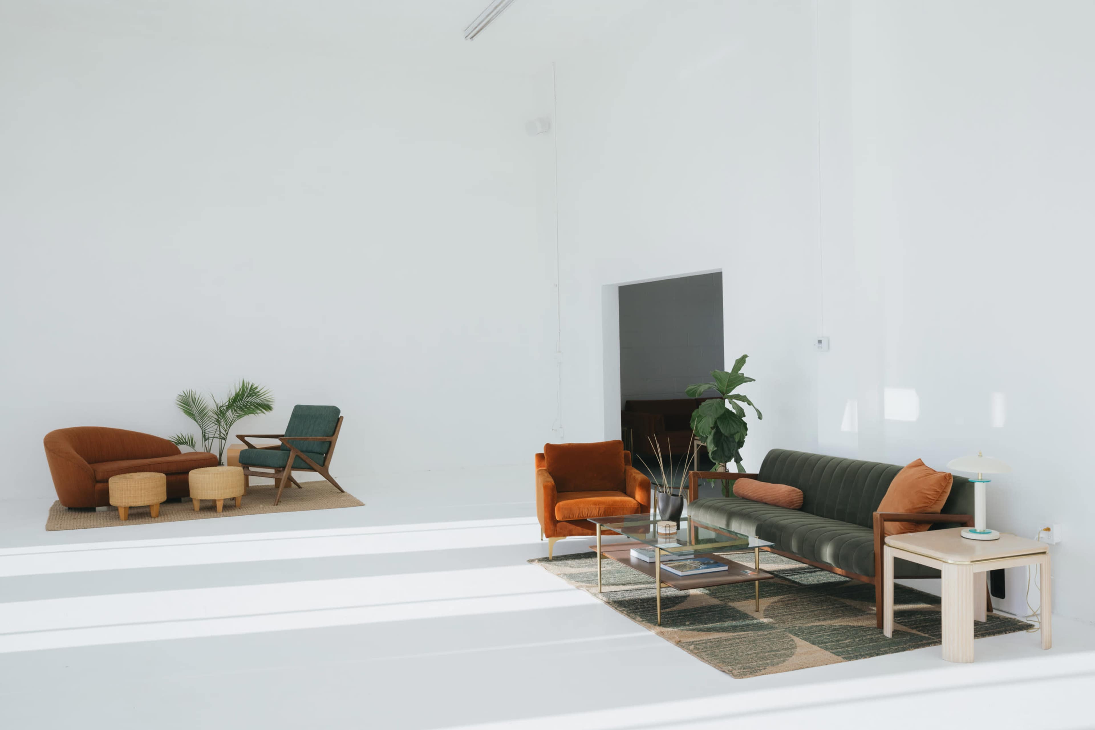 The image shows a minimalist living space with a green sofa, an orange chair, and a beige armchair on a light-colored floor, surrounded by plants and a coffee table.