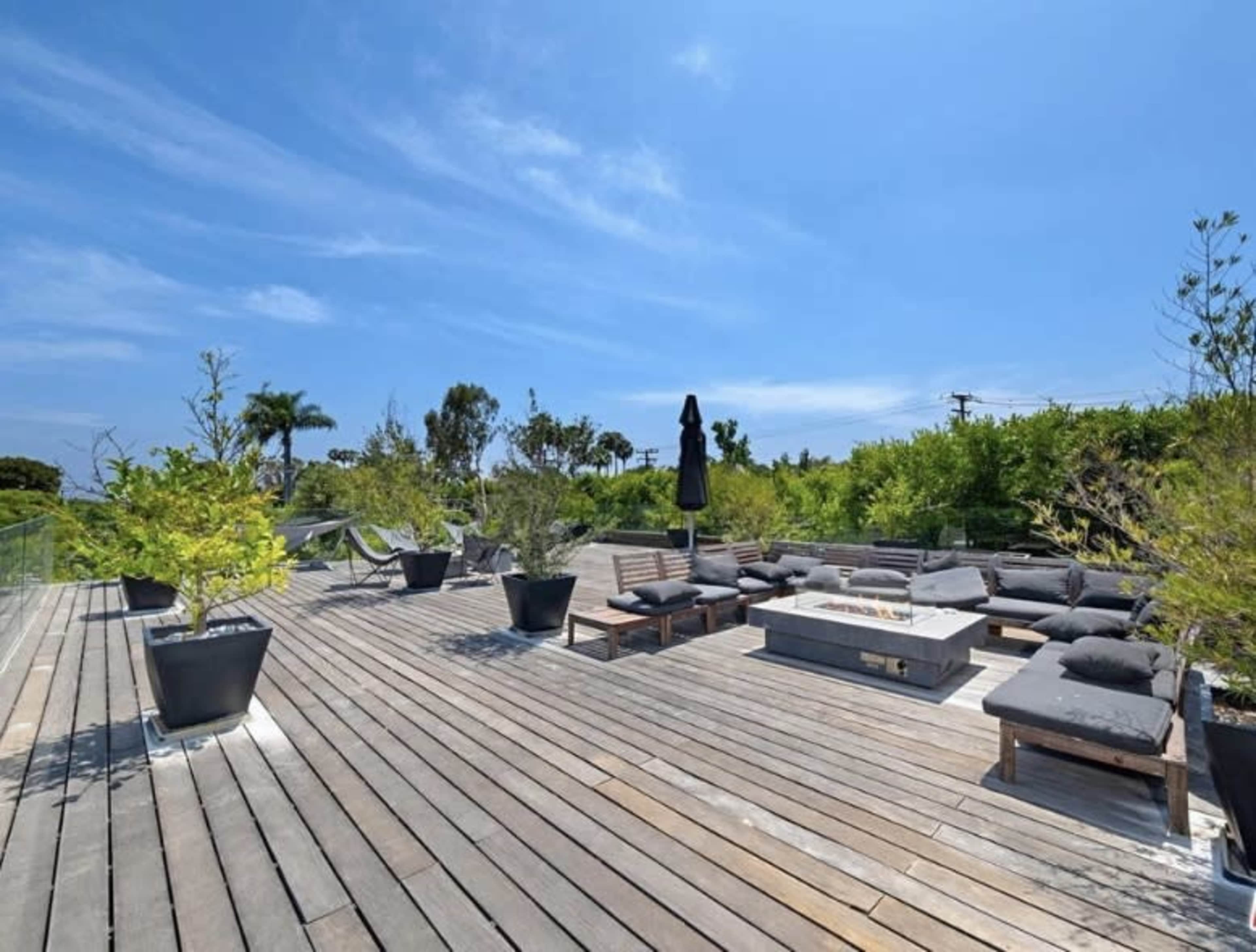 A spacious wooden deck features seating areas with cushions, potted plants, and a central fire pit under a clear blue sky.