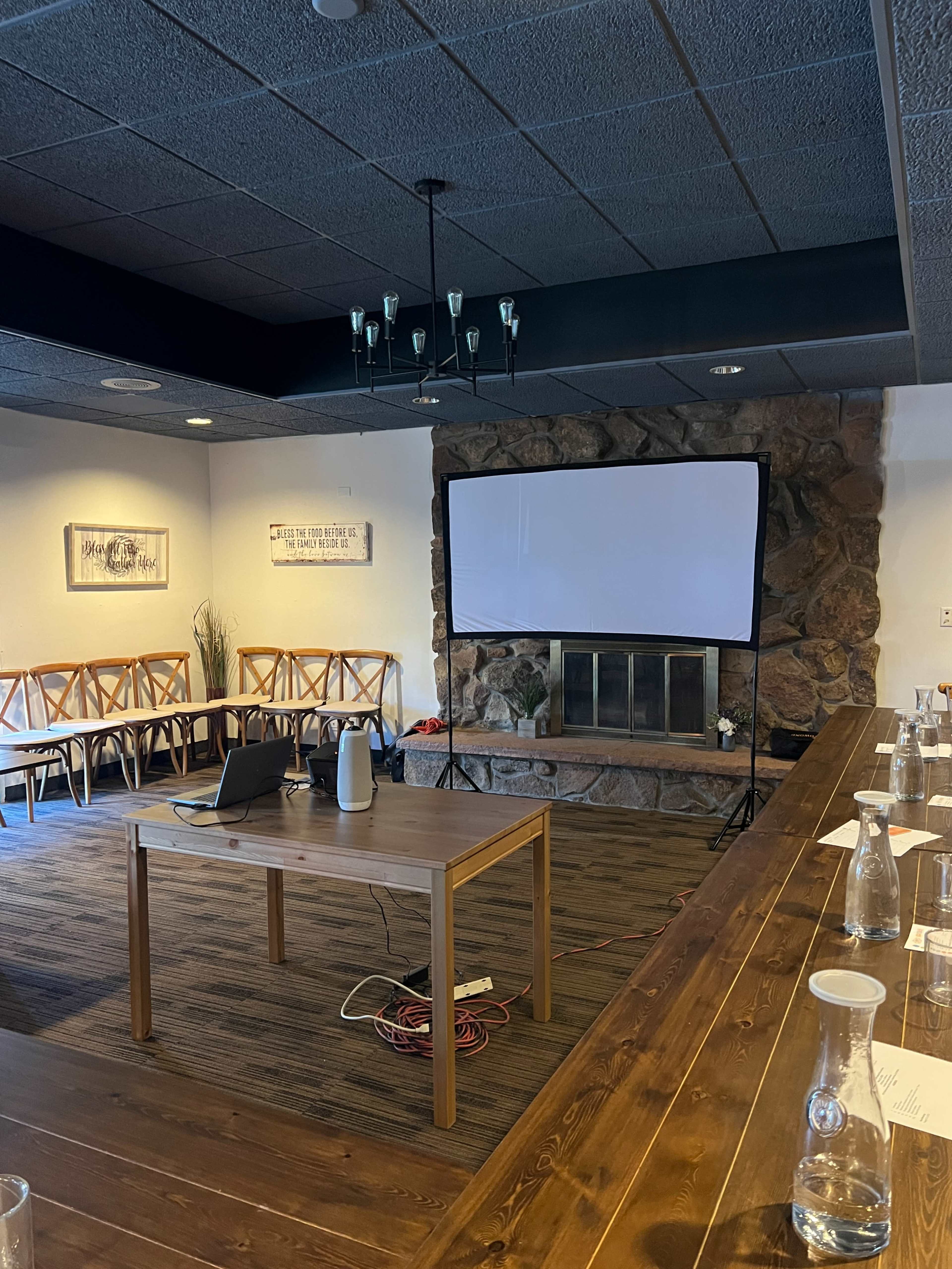 A meeting room with a wooden table, chairs arranged along the walls, a large screen on a stand, and a stone fireplace.
