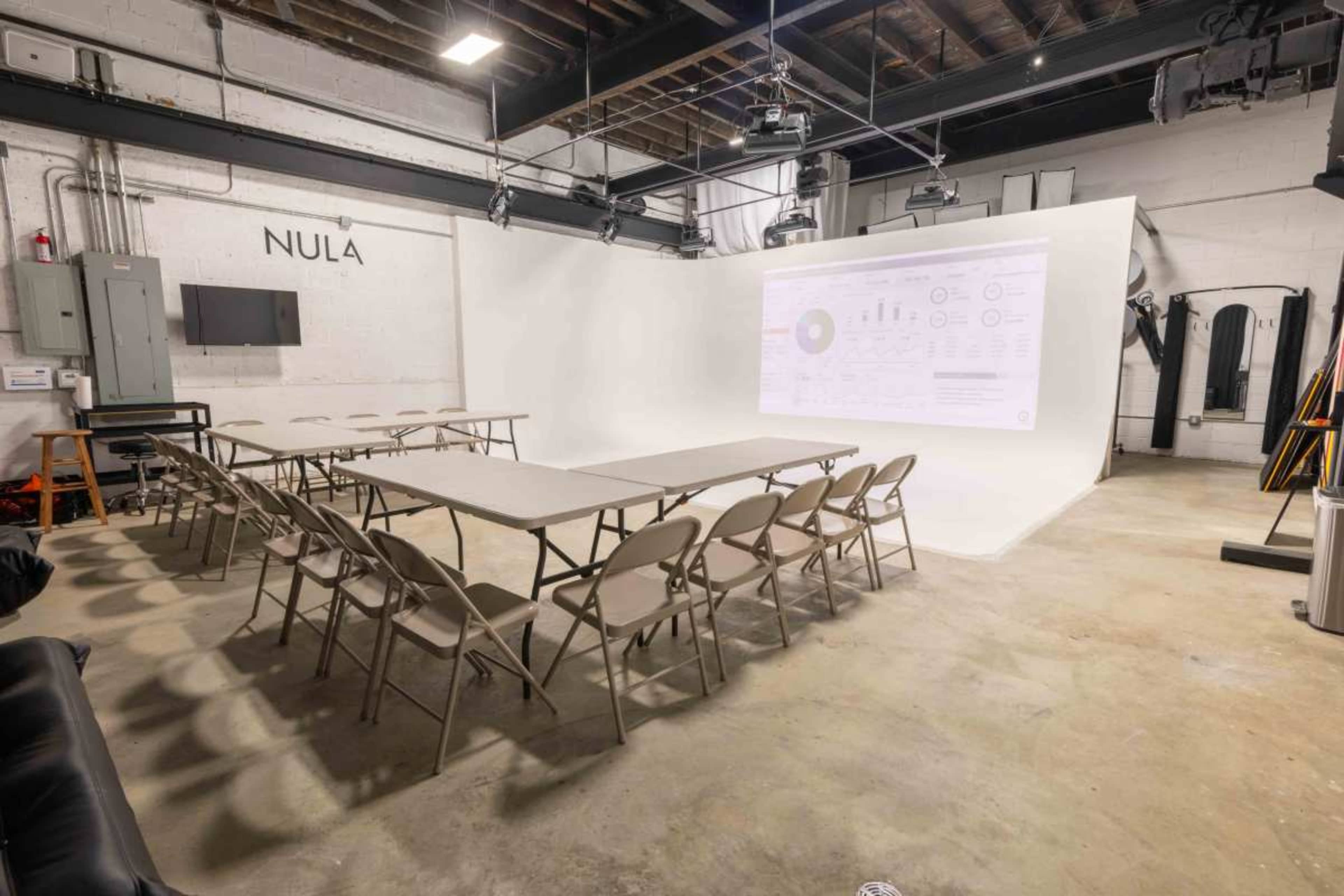 A minimalistic meeting space is set up with rows of folding tables and chairs facing a projector screen displaying data charts against a white background.