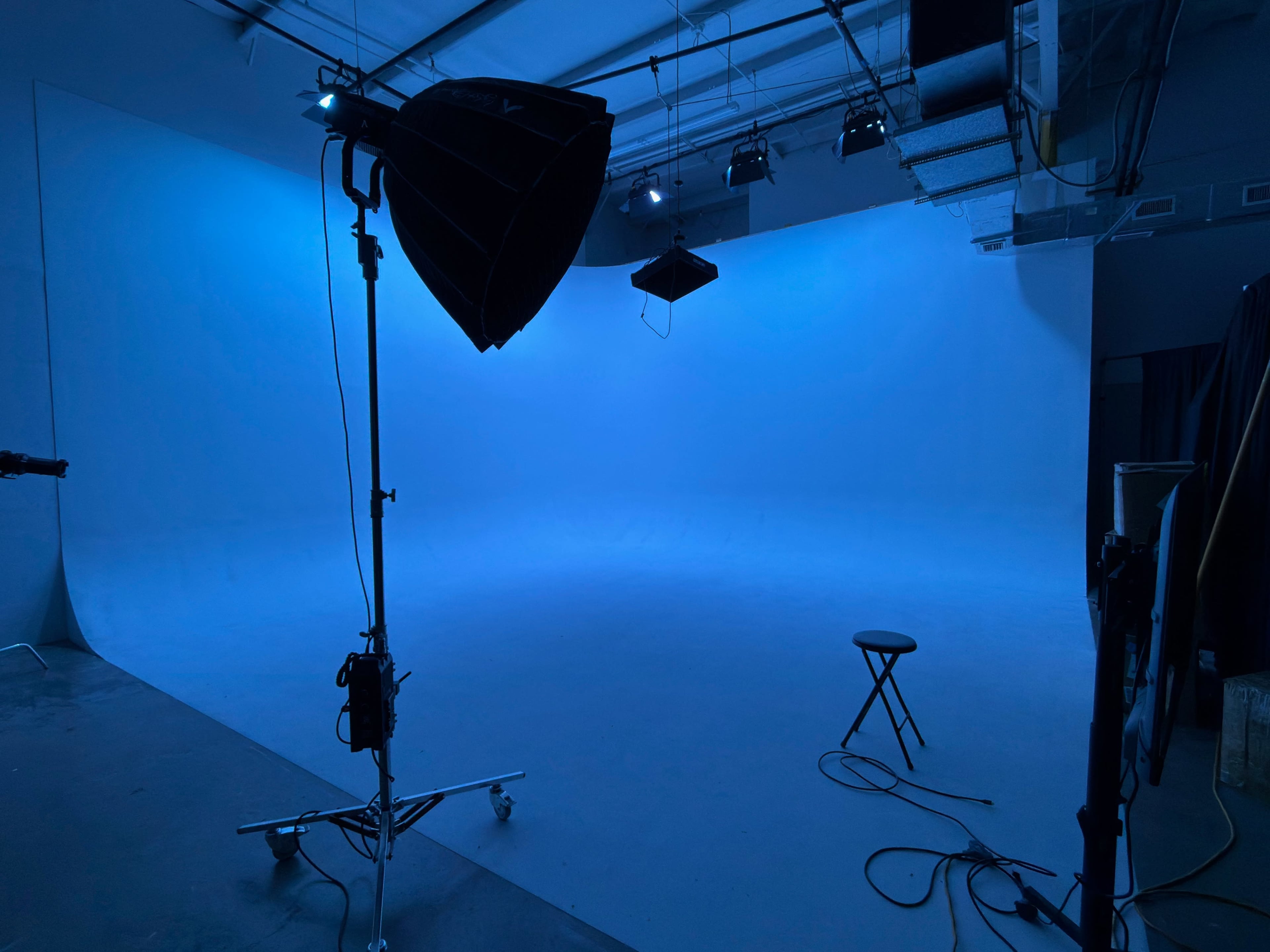 A photography studio features a blue backdrop, a lighting fixture on a stand, and a stool positioned in the center.