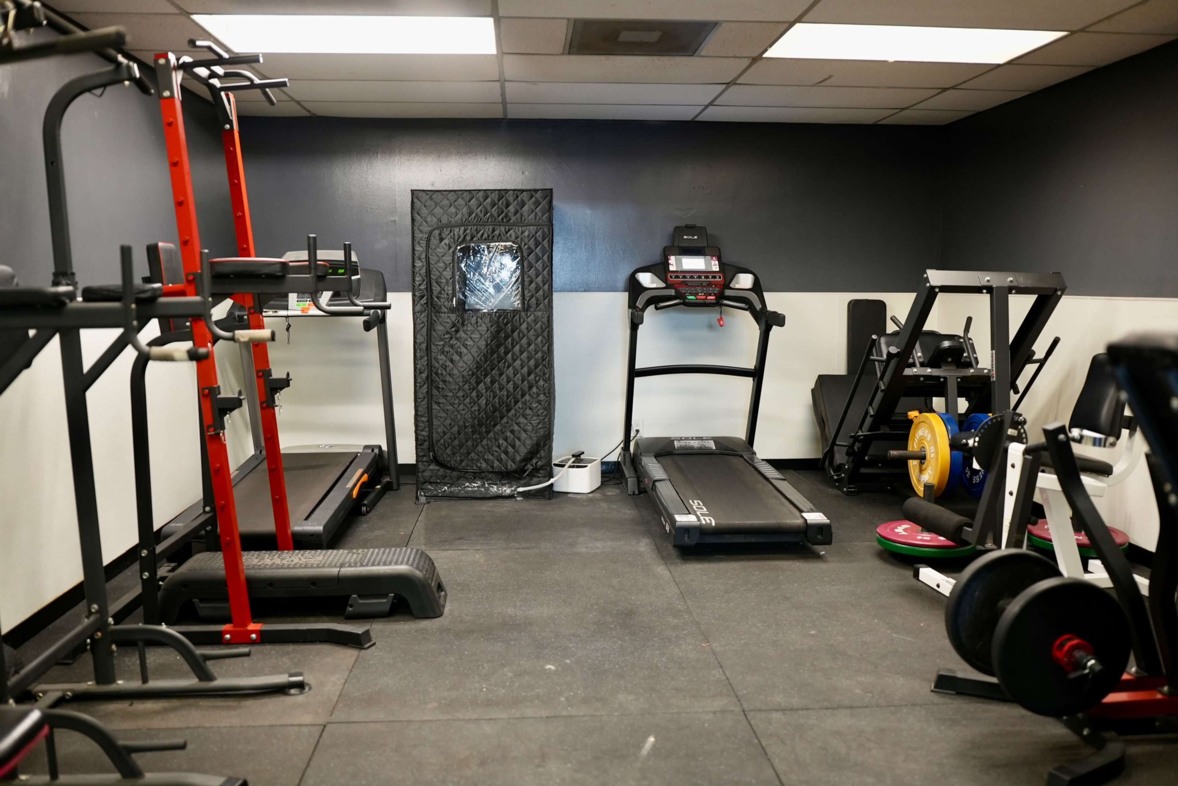 The image shows a small gym area with exercise equipment including a treadmill, weightlifting racks, and various weights on the floor.