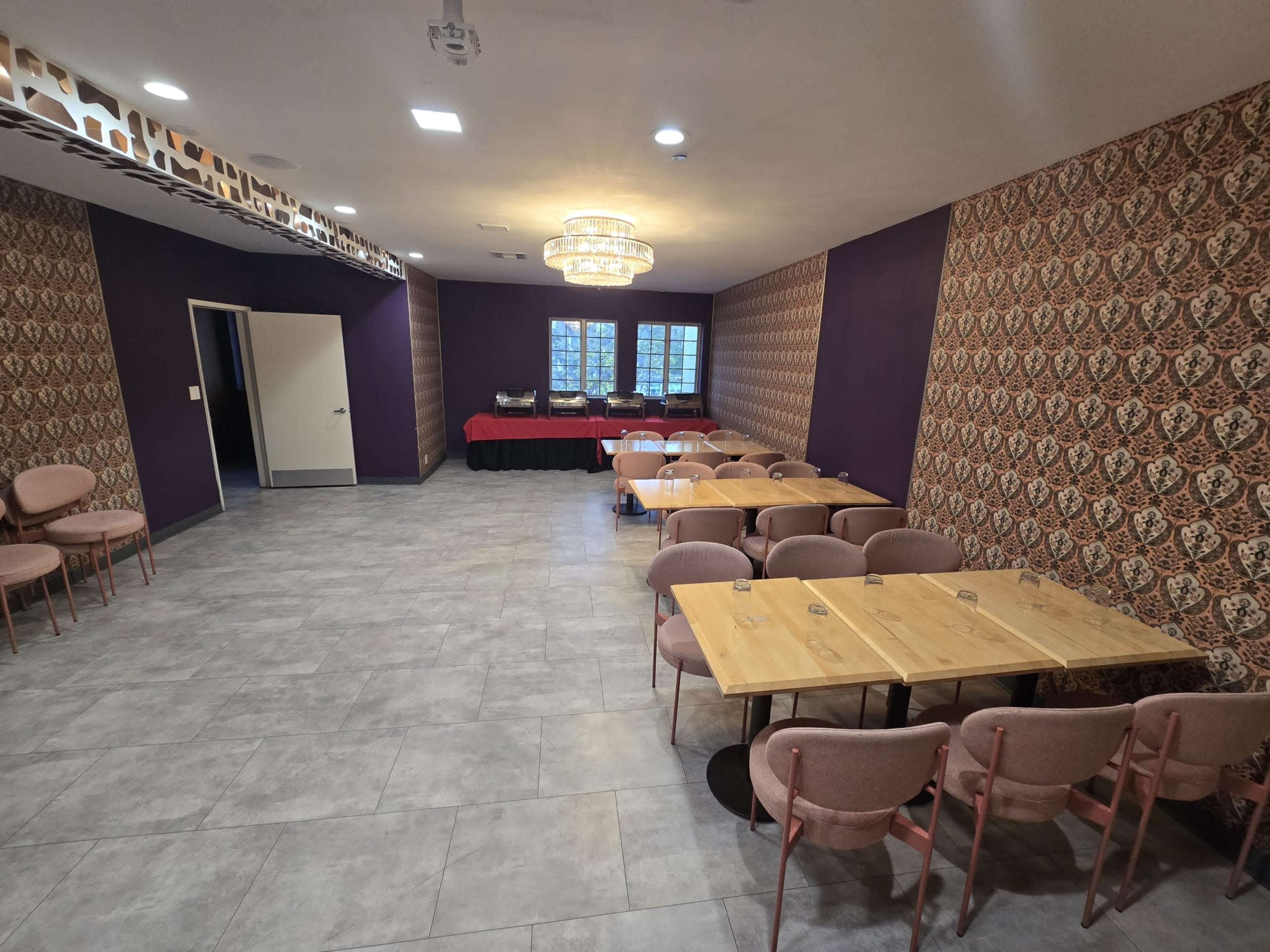 The image shows a modern room with patterned wallpaper, two wooden tables set for dining, and pink chairs arranged around them, with a buffet setup against one wall.