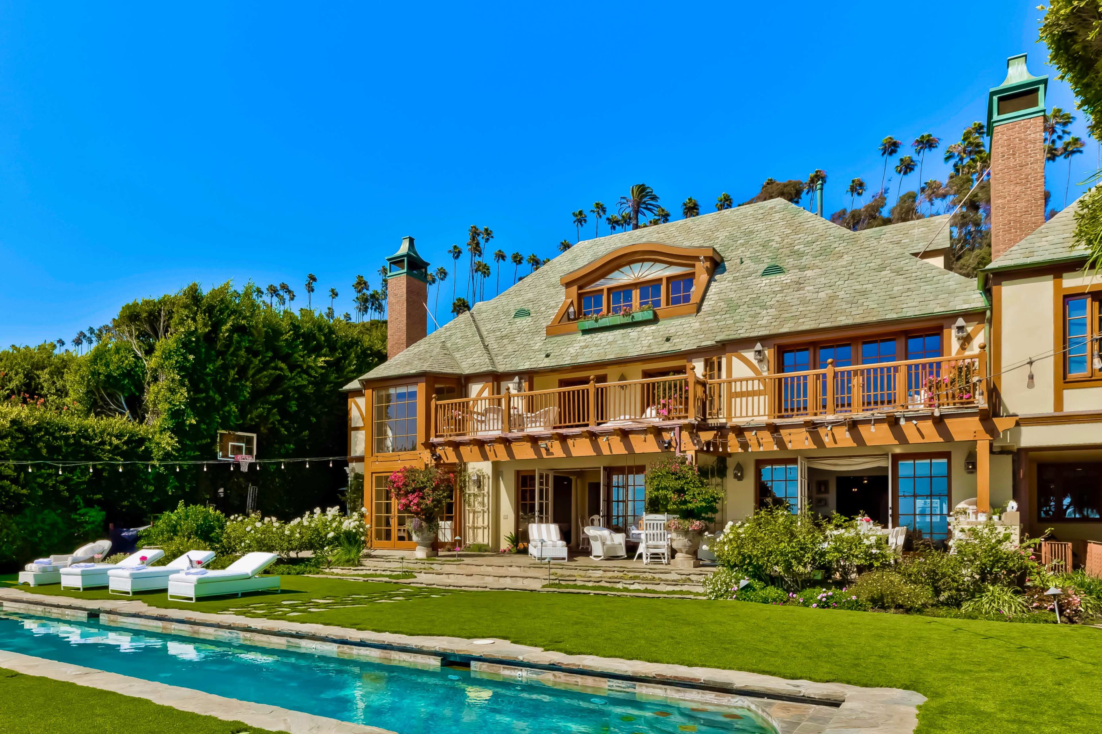 A large, two-story house with a stone patio and a swimming pool is surrounded by greenery and palm trees.