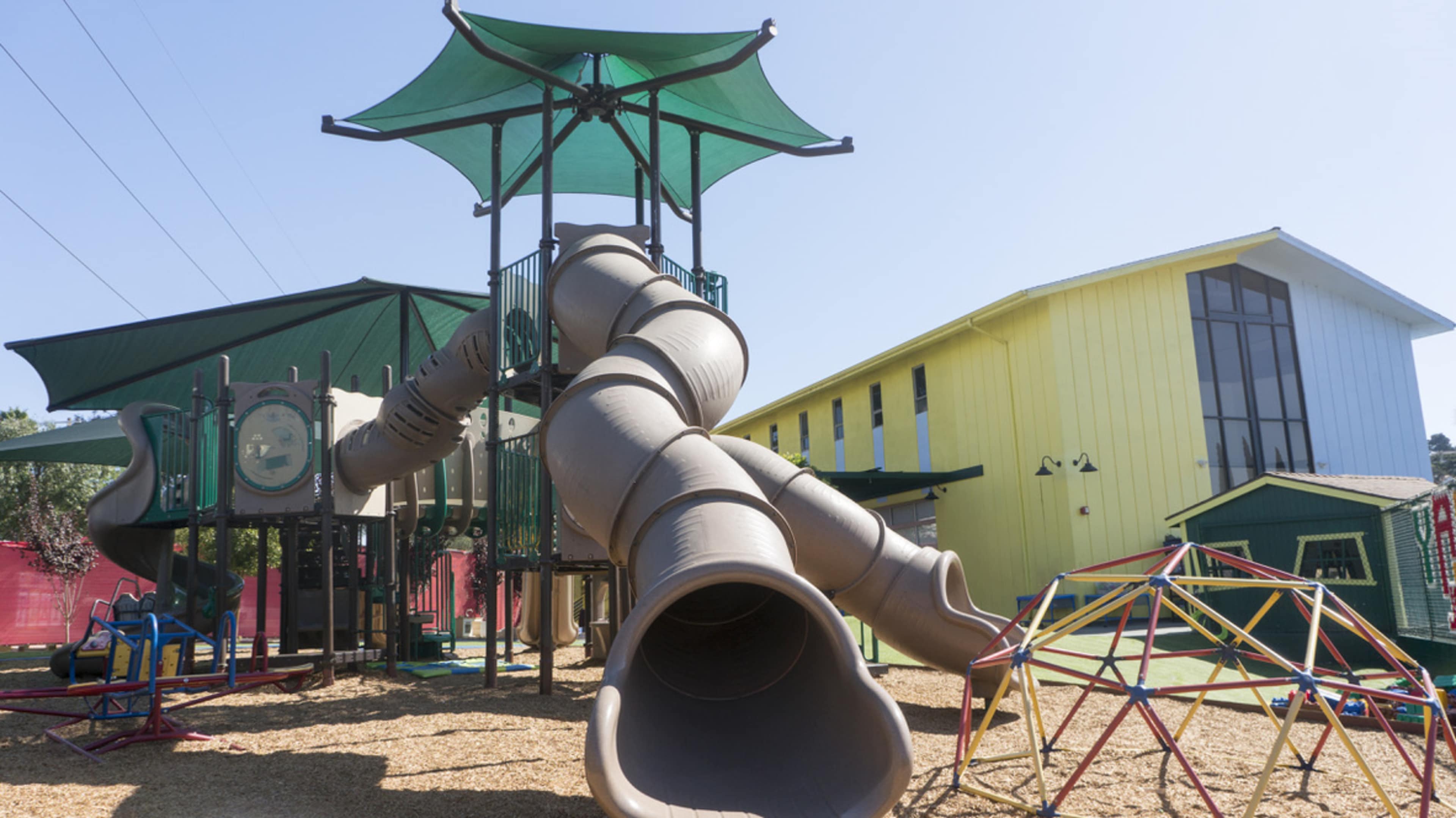 A large playground structure featuring multiple slides and a shaded area, with a yellow building in the background.