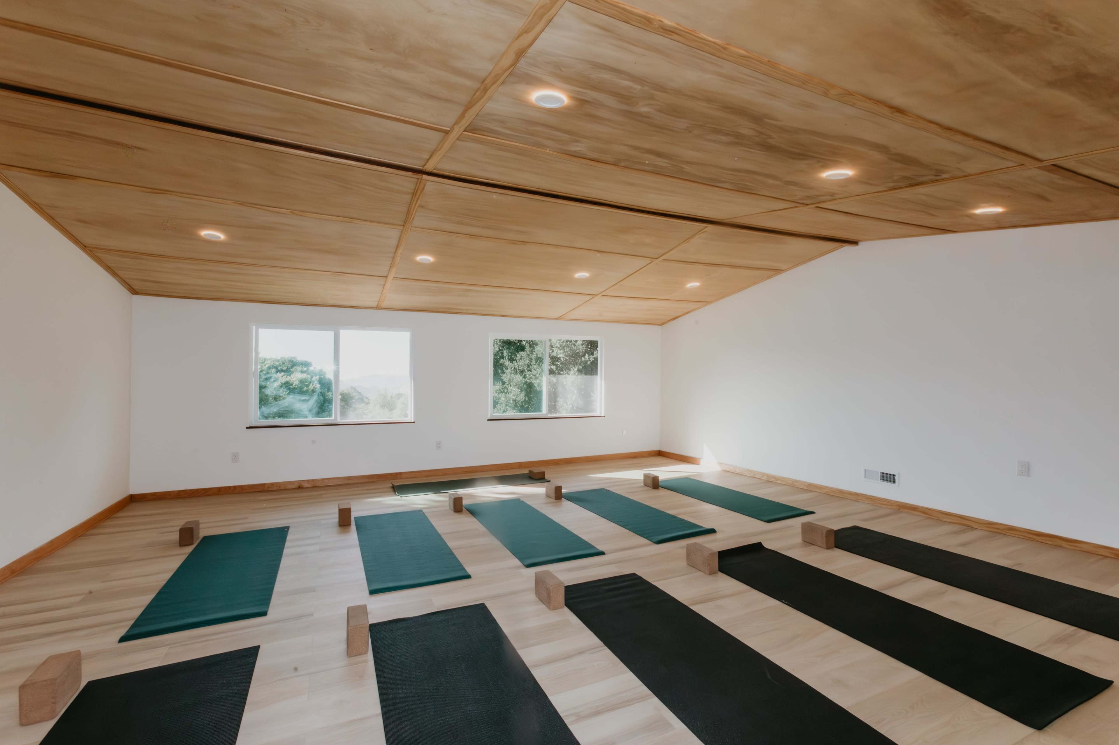 The image shows a spacious, well-lit yoga studio with wooden floors, arranged yoga mats, and blocks laid out for practice.