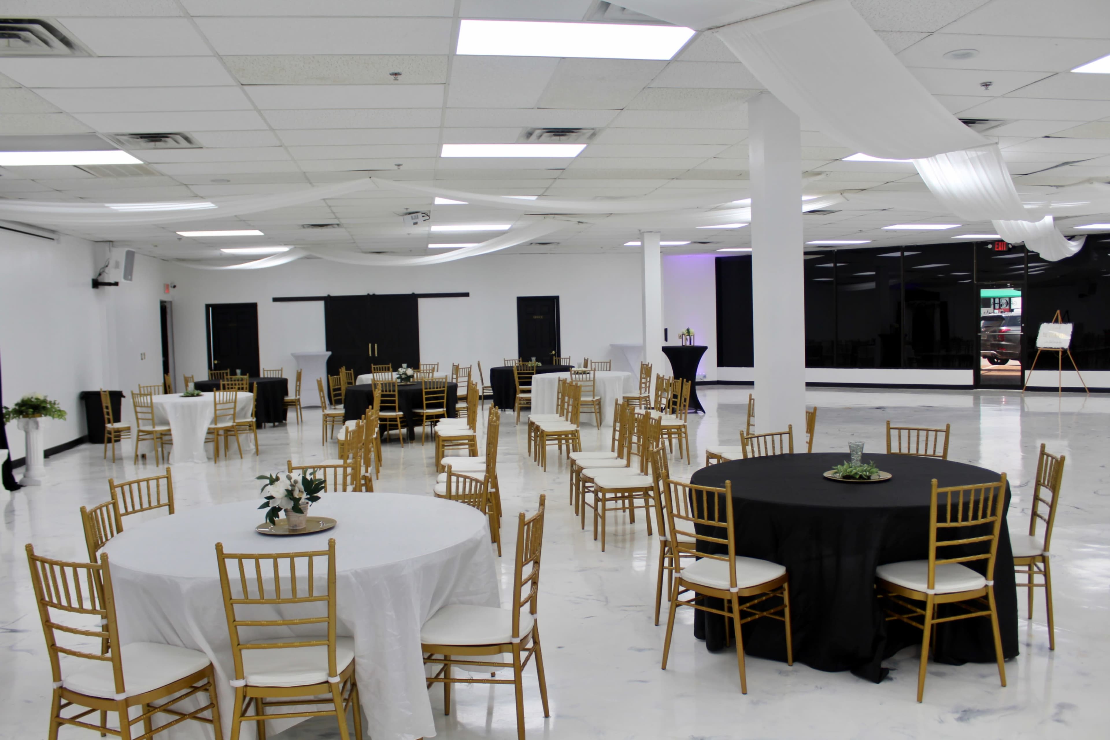 A spacious, empty event hall features round tables with black and white tablecloths arranged around the room under white draped fabric.
