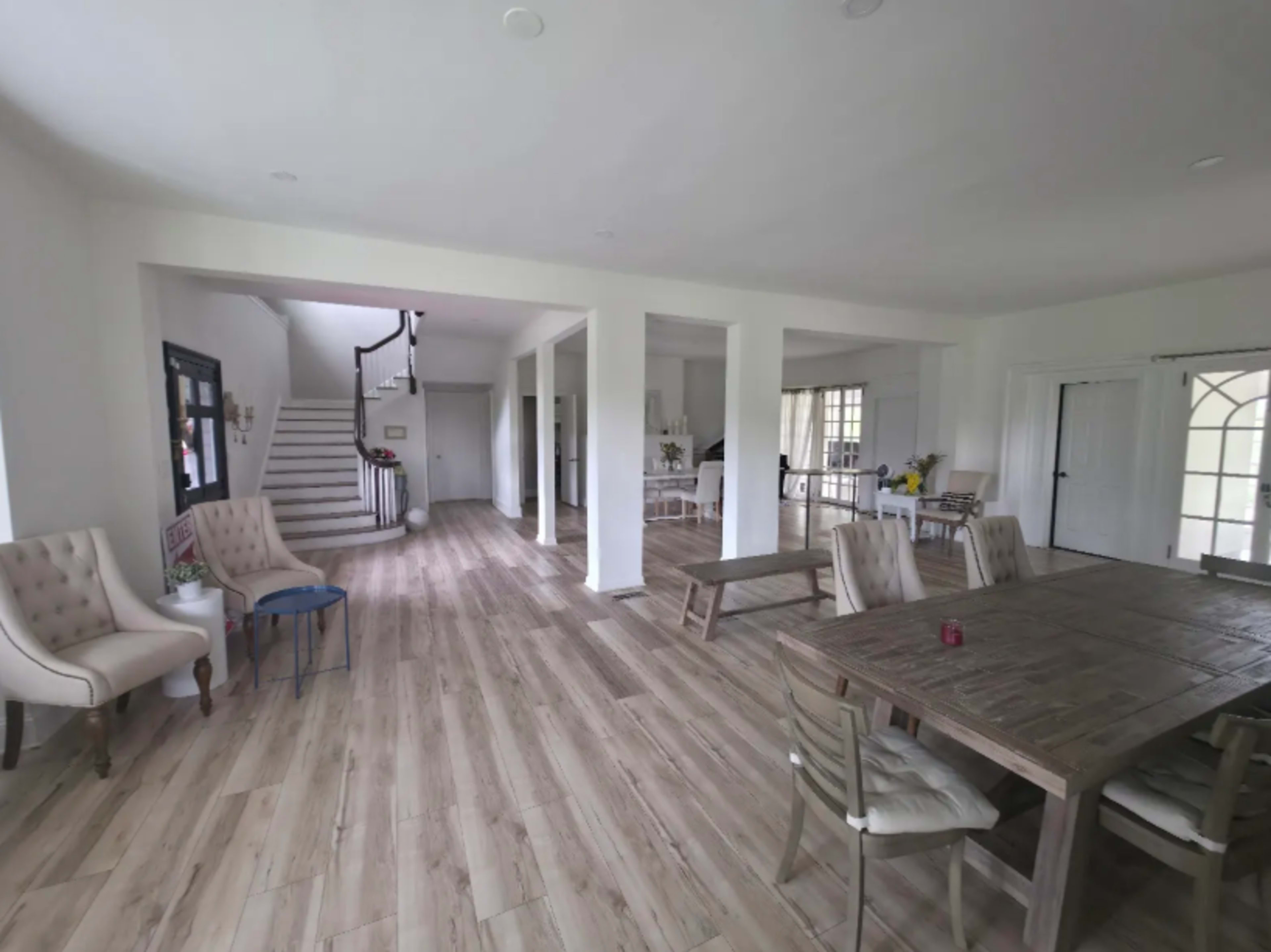 The image shows an open, airy interior space with wooden flooring, featuring a dining table, chairs, and a staircase in the background.