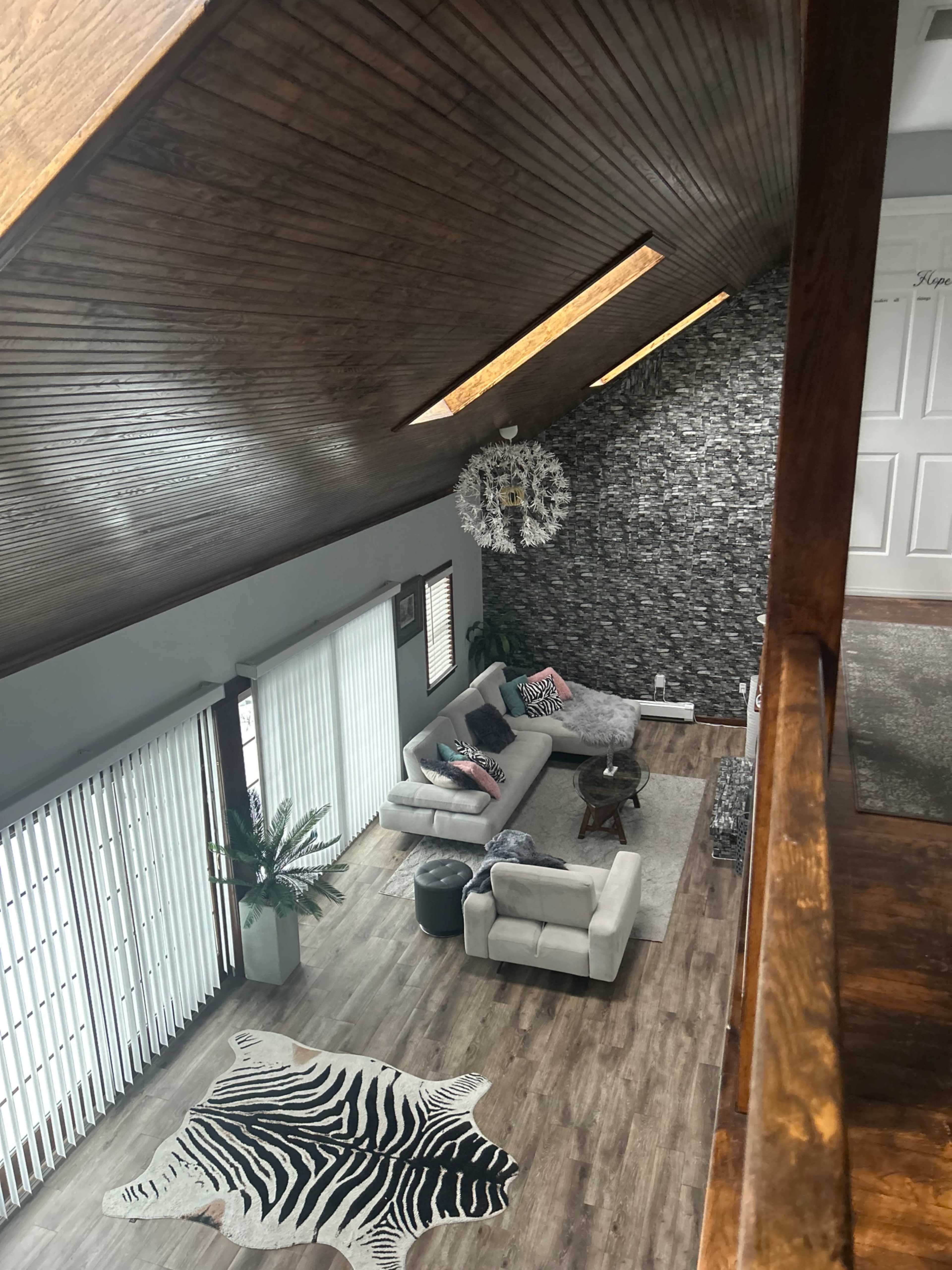 The image shows a modern living room with a sectional sofa, a patterned rug, and large windows that let in natural light, all under a wooden ceiling with a decorative wall.