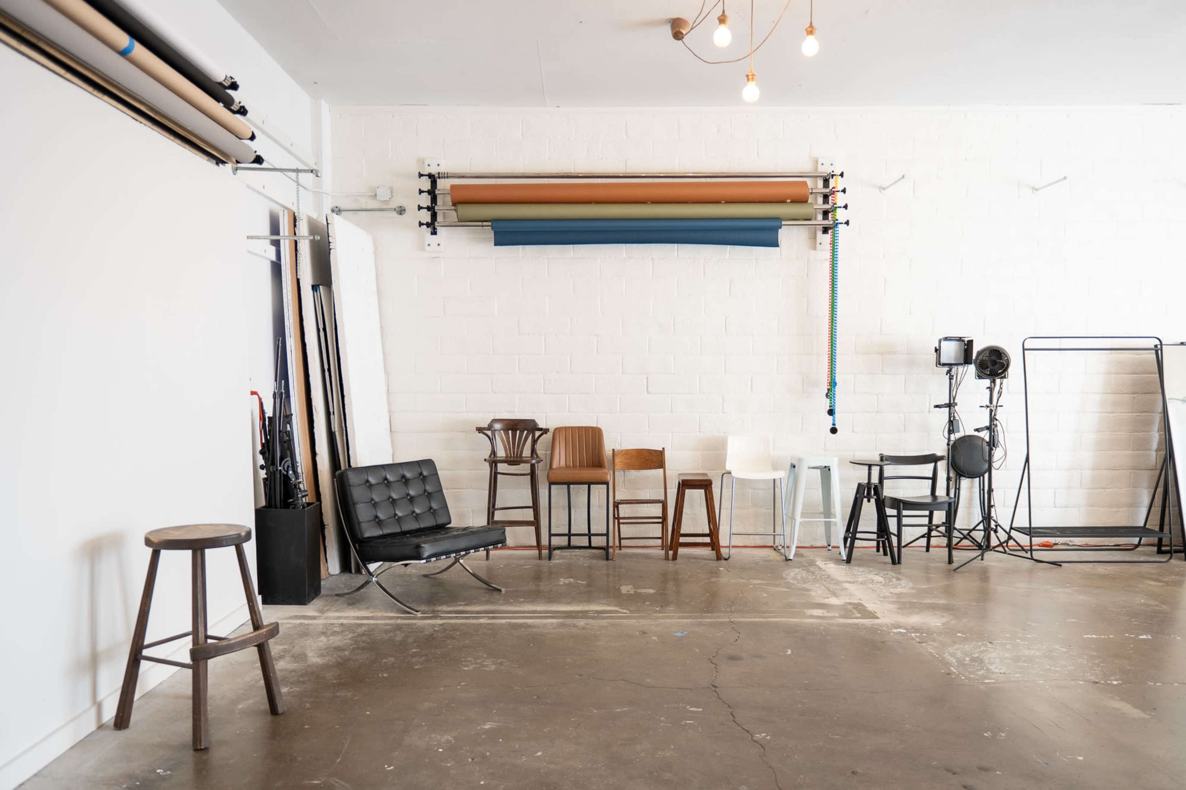 The image shows an interior space with a mix of wooden and metal chairs arranged against a white wall, along with rolls of background paper and single stools.