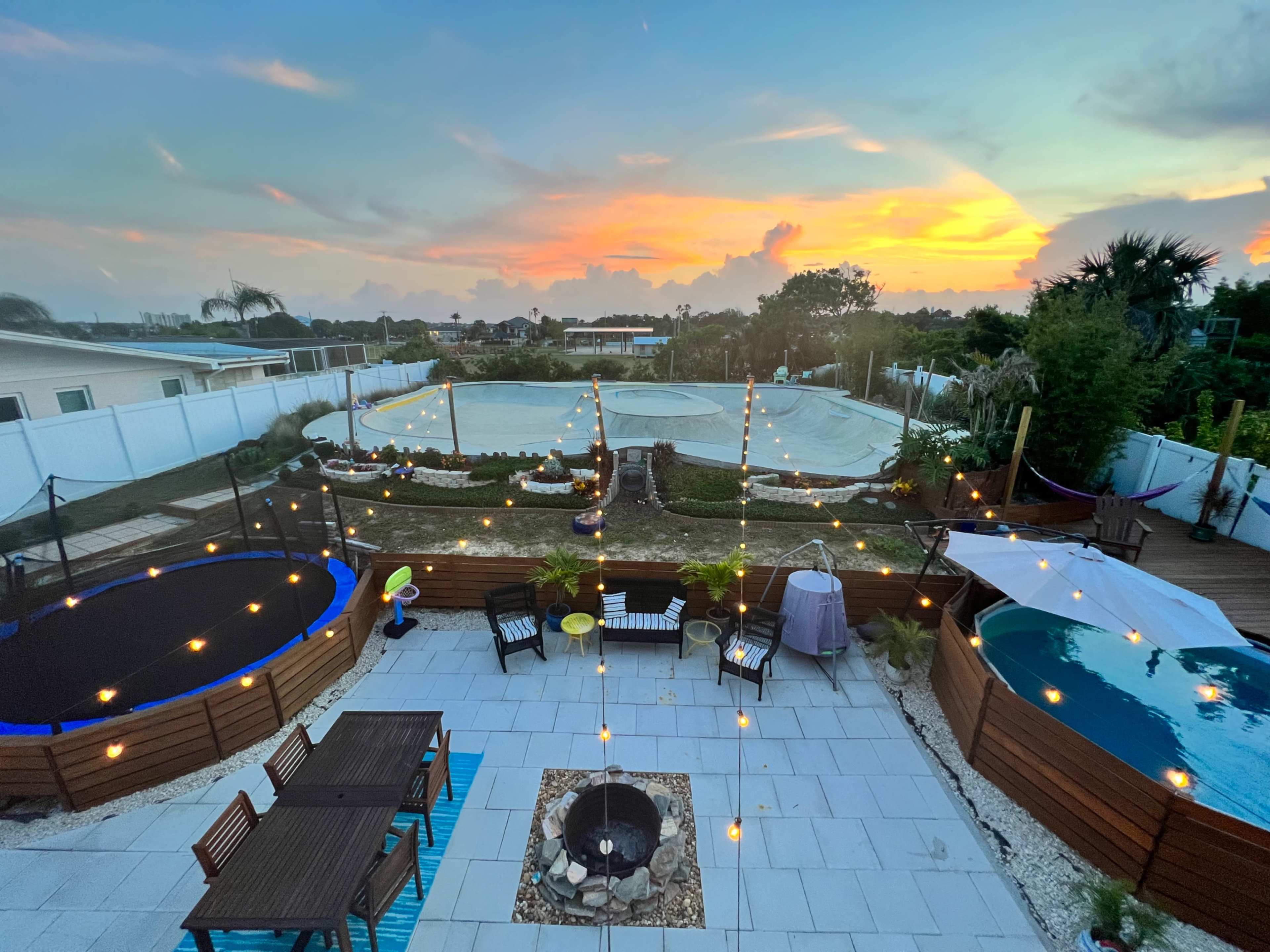 The image shows a backyard scene featuring a sunset, with multiple pools and trampolines surrounded by string lights and patio furniture.