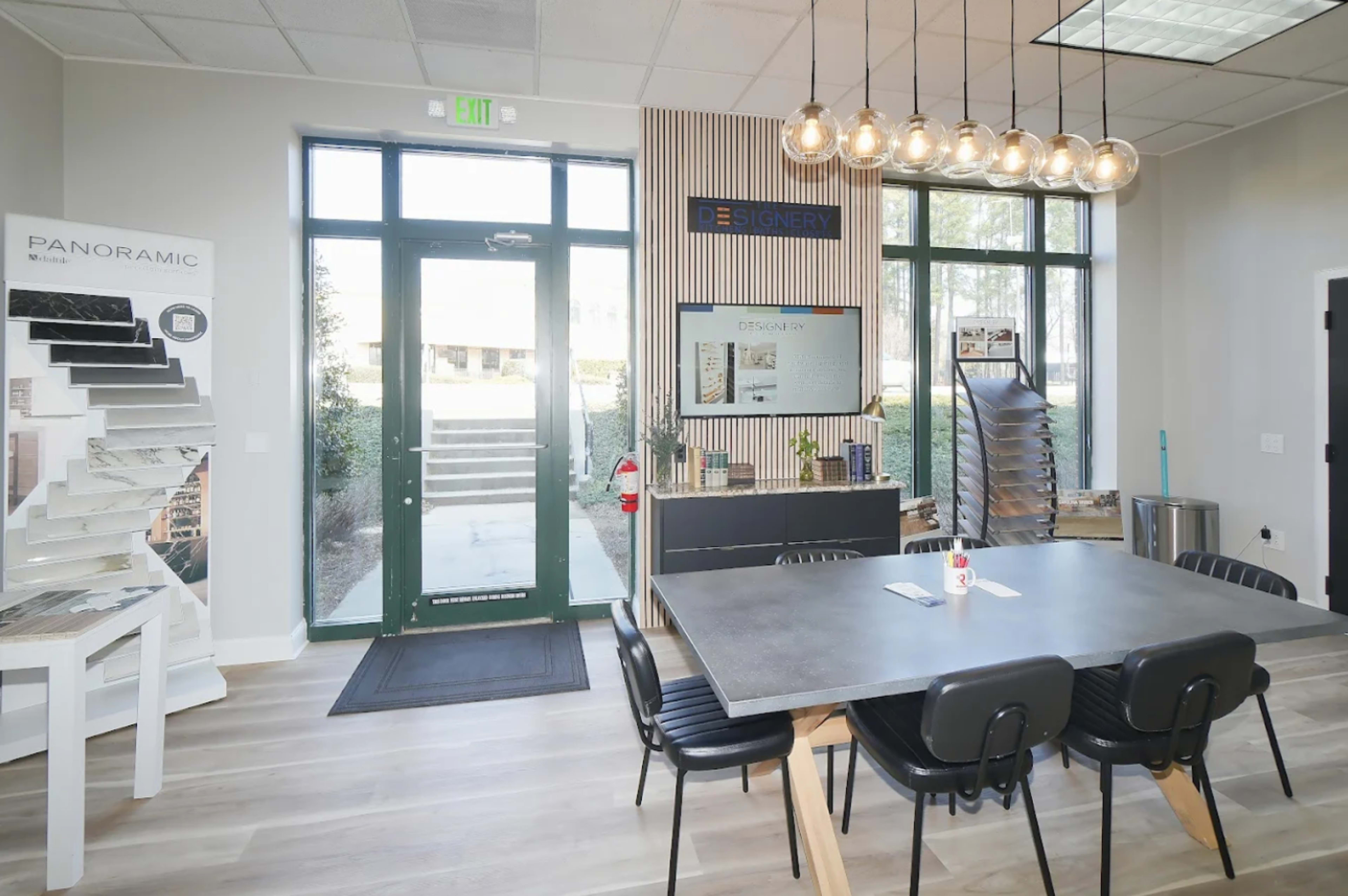 The image shows a modern workspace featuring a large table surrounded by chairs, with large windows and a door leading outside, along with design samples displayed on the walls.