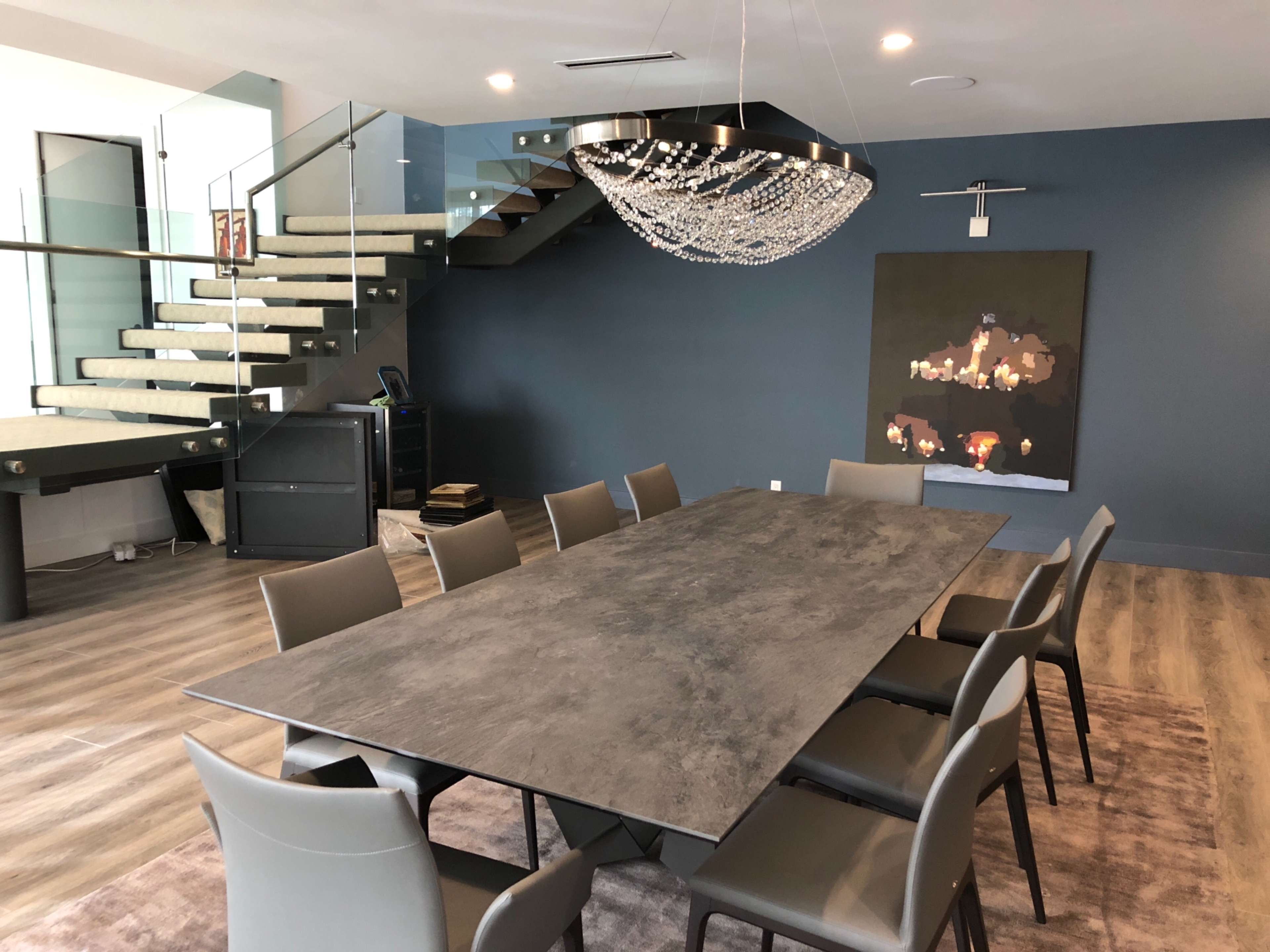 A modern dining area features a long rectangular table surrounded by eight chairs, with a staircase in the background and a chandelier overhead.