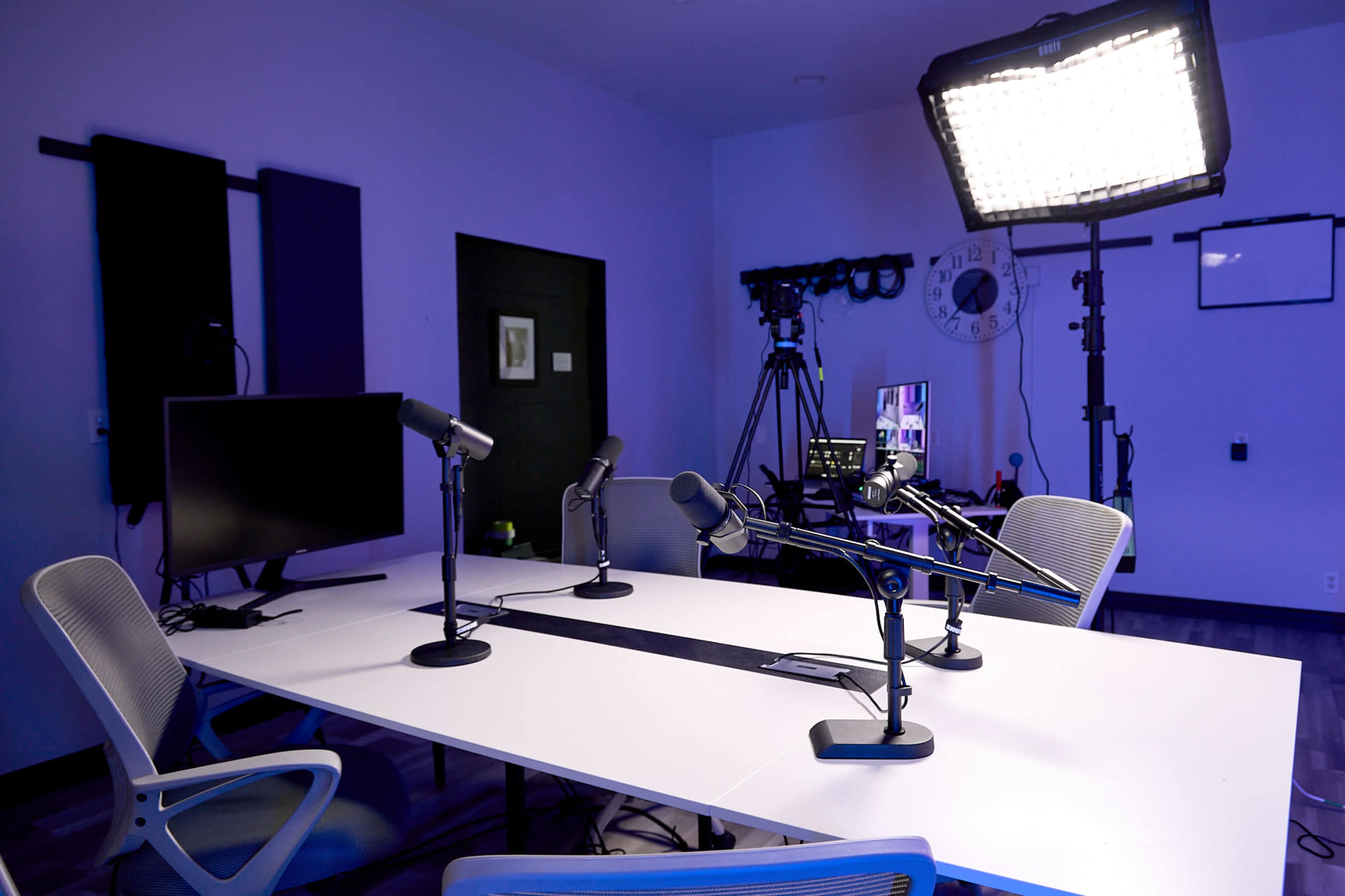 A podcast recording studio with a white table, four microphones, a monitor, and studio lighting.