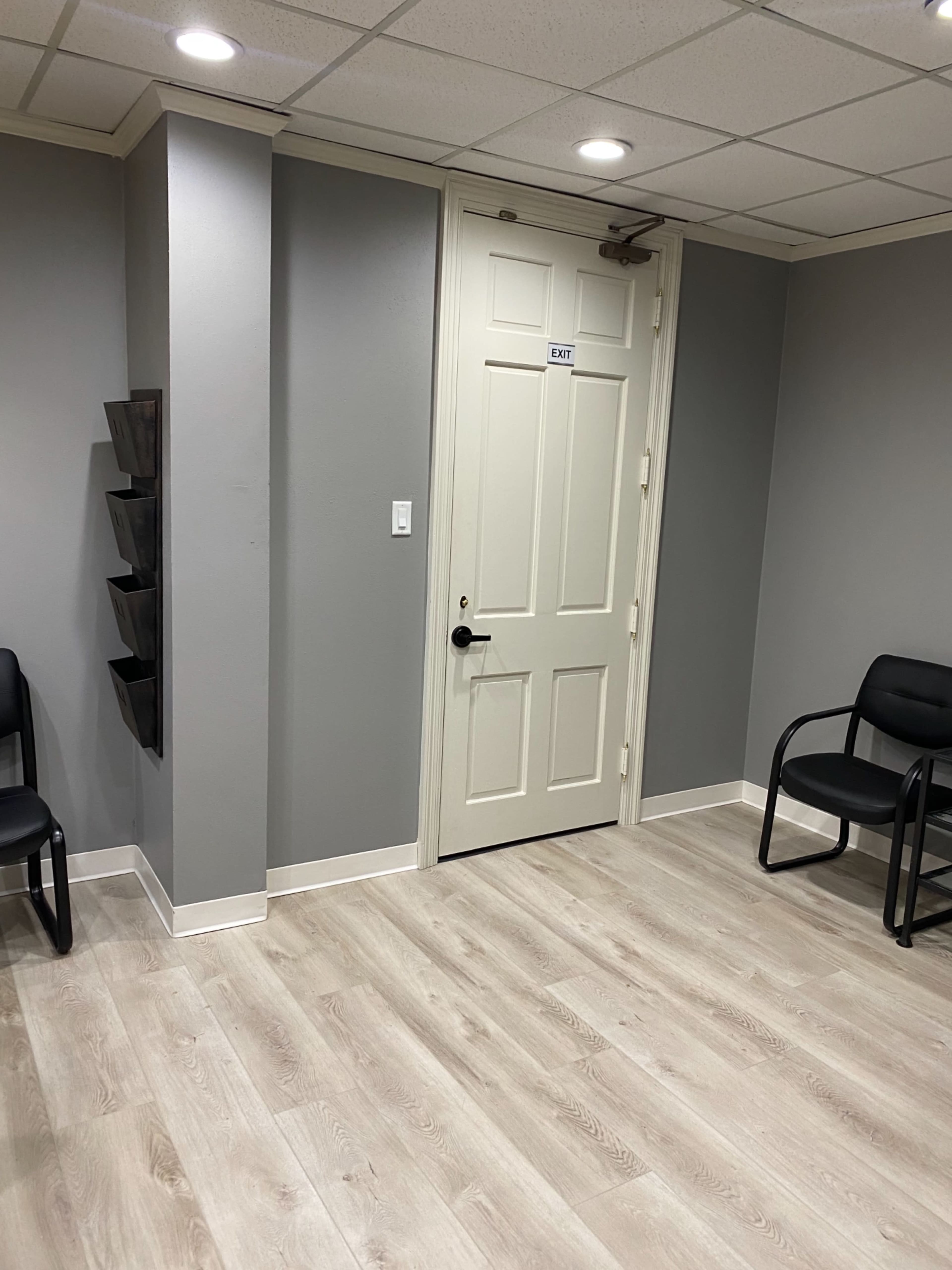 The image shows a waiting room with a door labeled "EXIT," gray walls, light-colored flooring, and several black chairs.