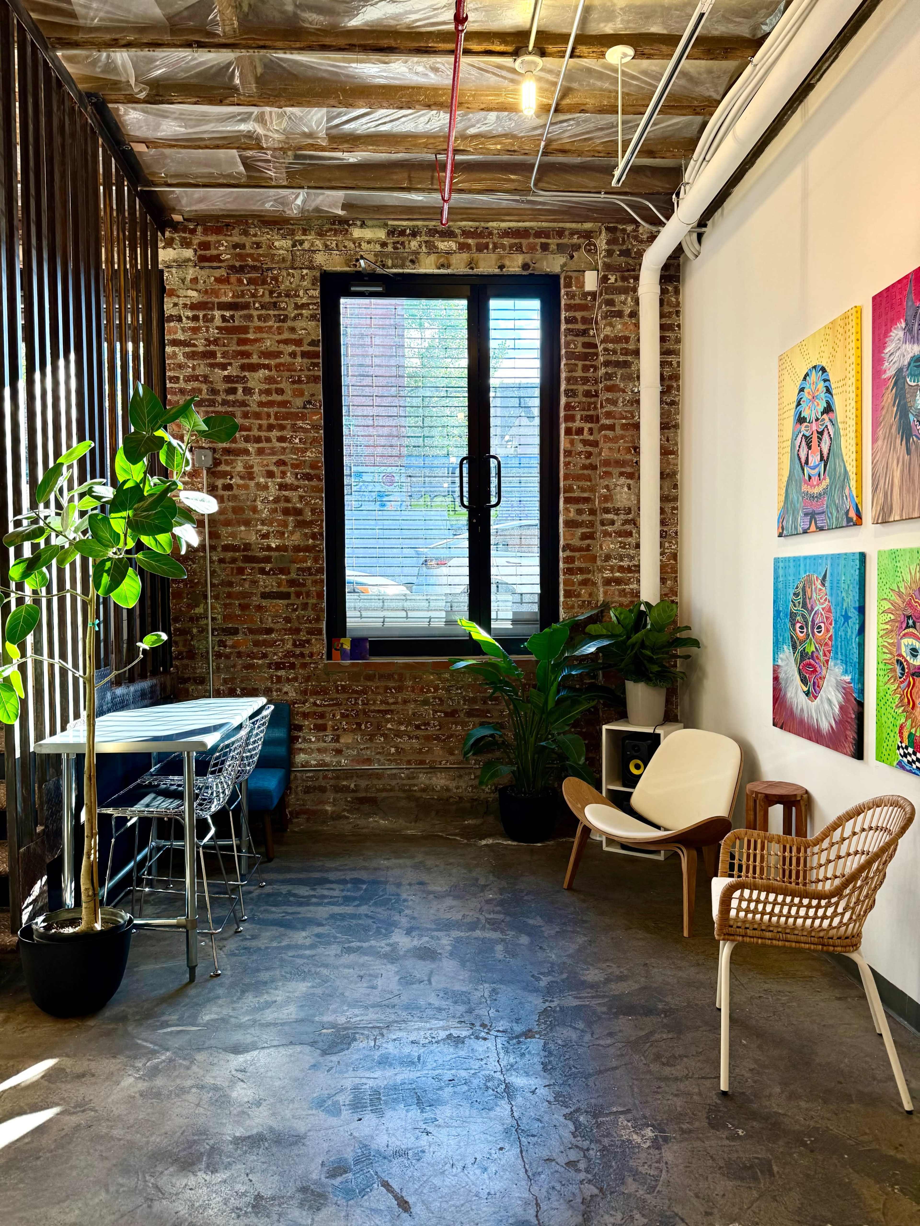 The image shows a stylish room with exposed brick walls, a window, artwork on the wall, and a mix of modern furniture including chairs and a table.