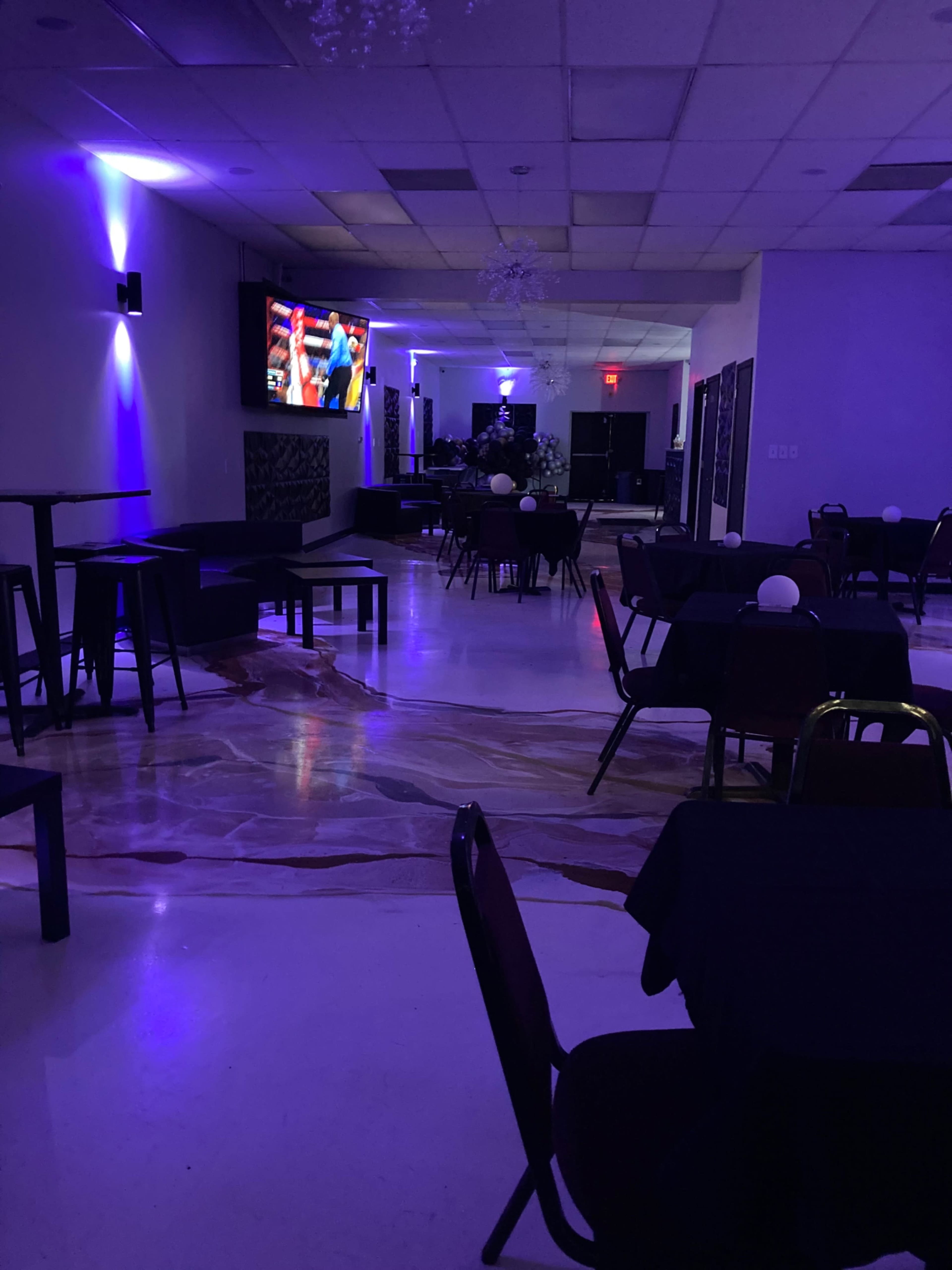 An empty restaurant with dark tables and chairs, illuminated by purple lighting and featuring a television screen displaying a sports event.