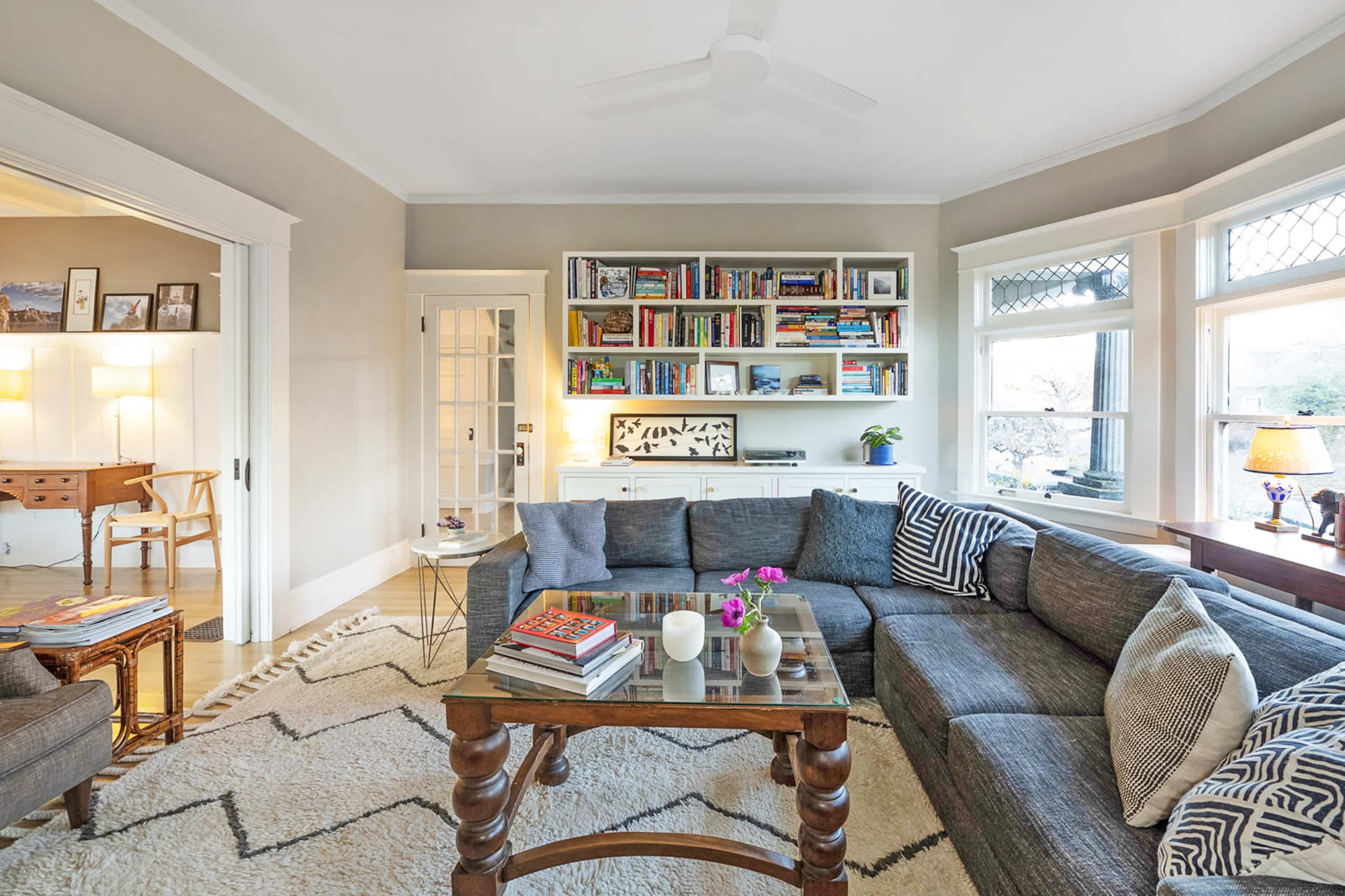 A cozy living room with a large sectional sofa, a coffee table stacked with books, and shelves filled with various titles, all illuminated by natural light from large windows.