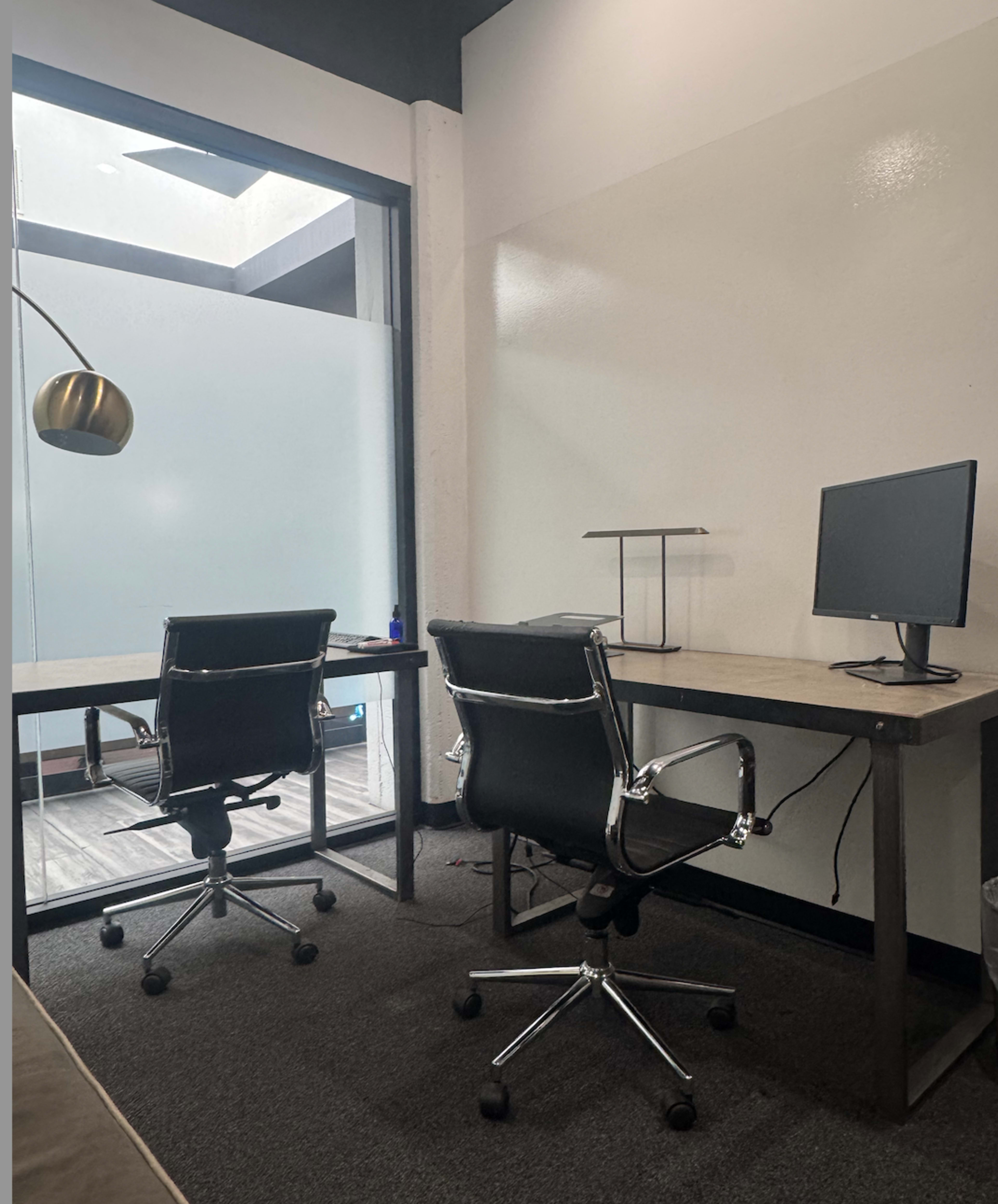 The image shows a modern office space with two black chairs positioned at desks, one of which has a computer monitor on it, and a glass partition allowing light to enter.