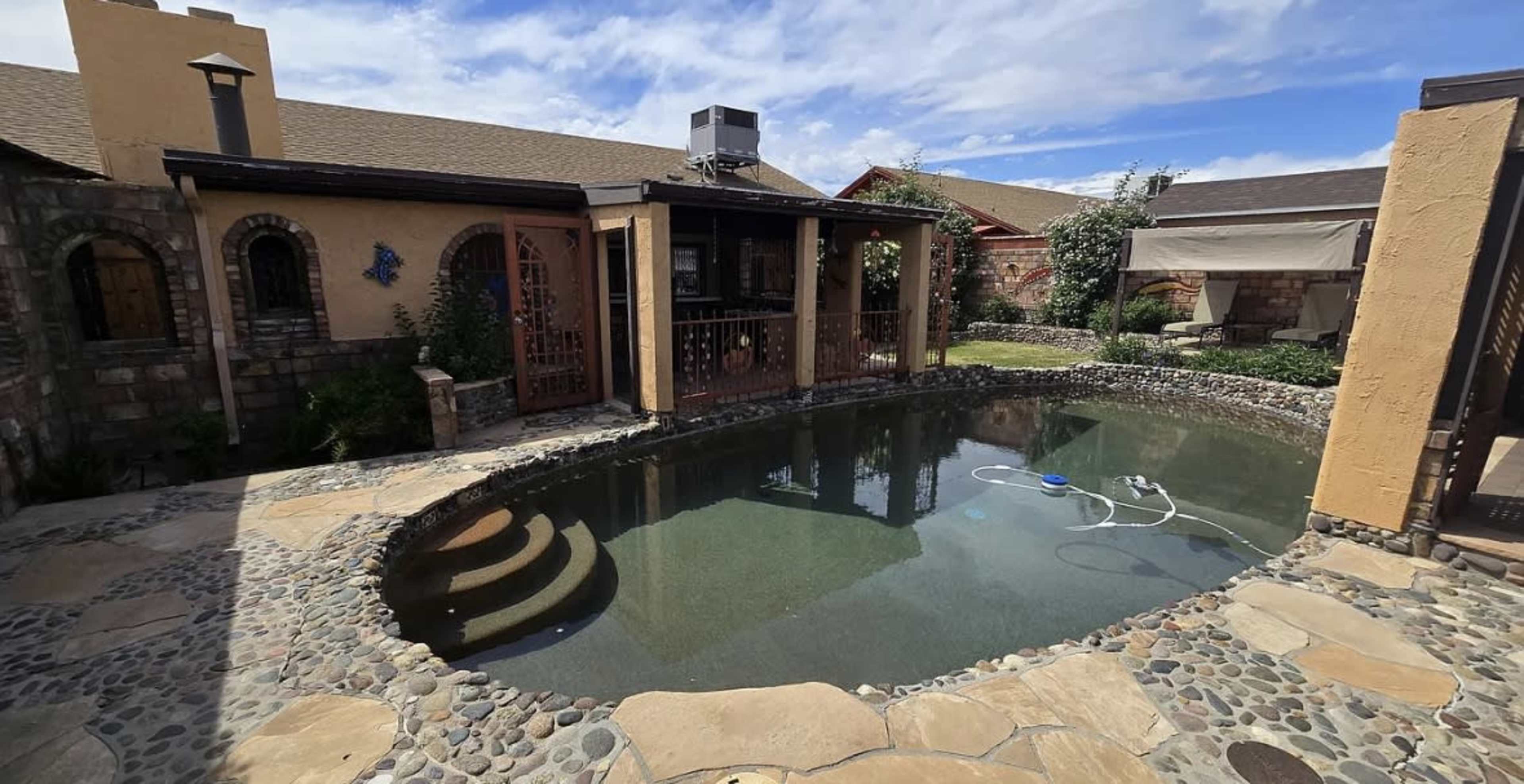 The image shows a landscaped backyard featuring a stone-lined pool surrounded by a patio and a house in the background.