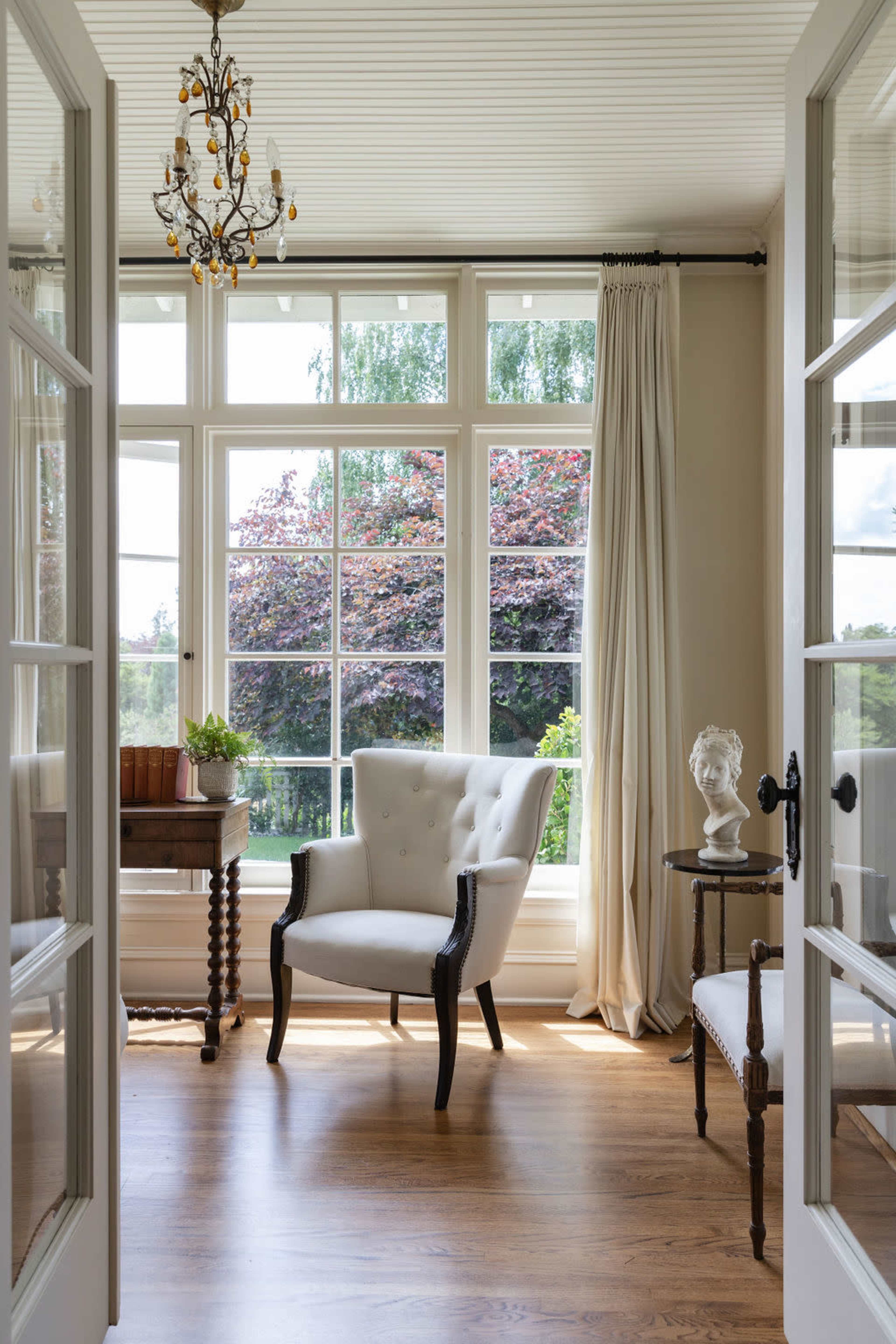 A bright, airy room features a white upholstered armchair, a small side table, and large windows that offer a view of lush greenery outside.