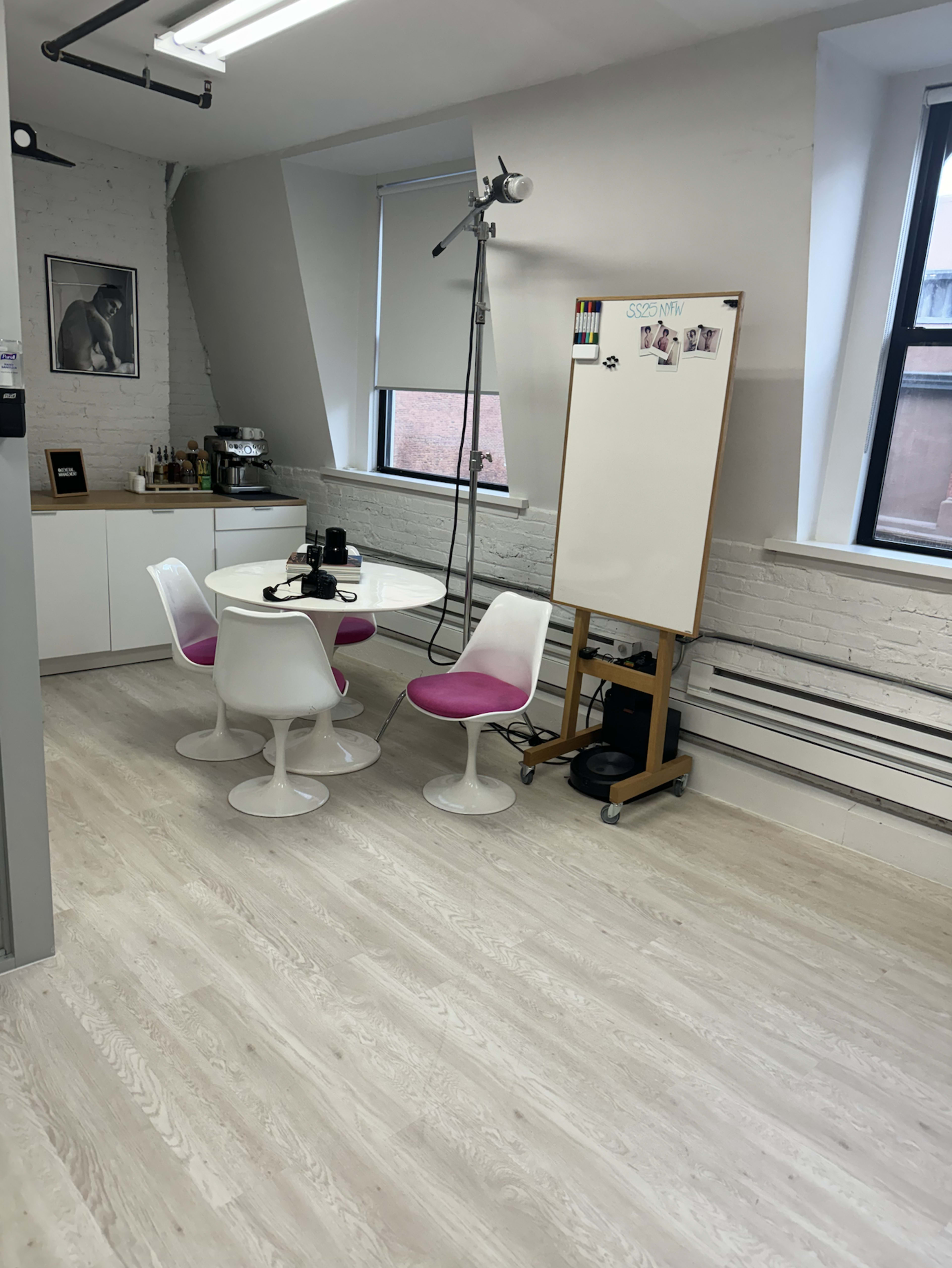 The image shows a bright, modern office space with a round table and four chairs, a whiteboard on an easel, and a small coffee station against the wall.