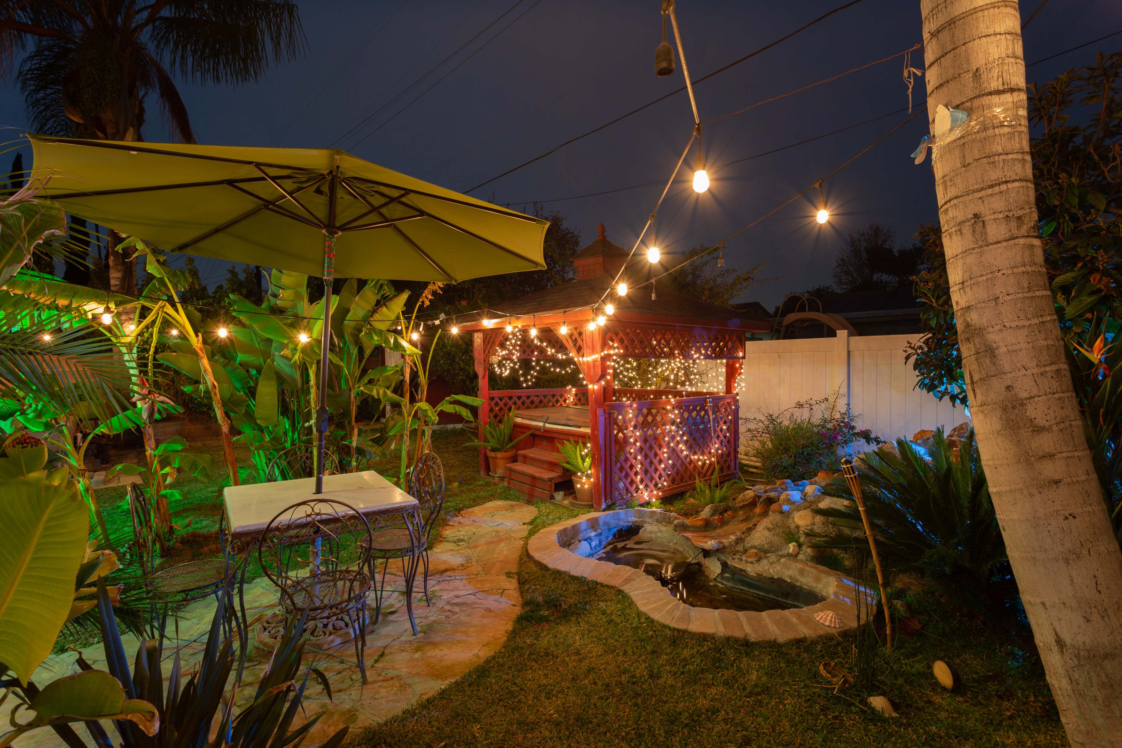 A backyard garden at night, featuring a patio with a table and chairs, a gazebo adorned with string lights, and a small water feature surrounded by tropical plants.