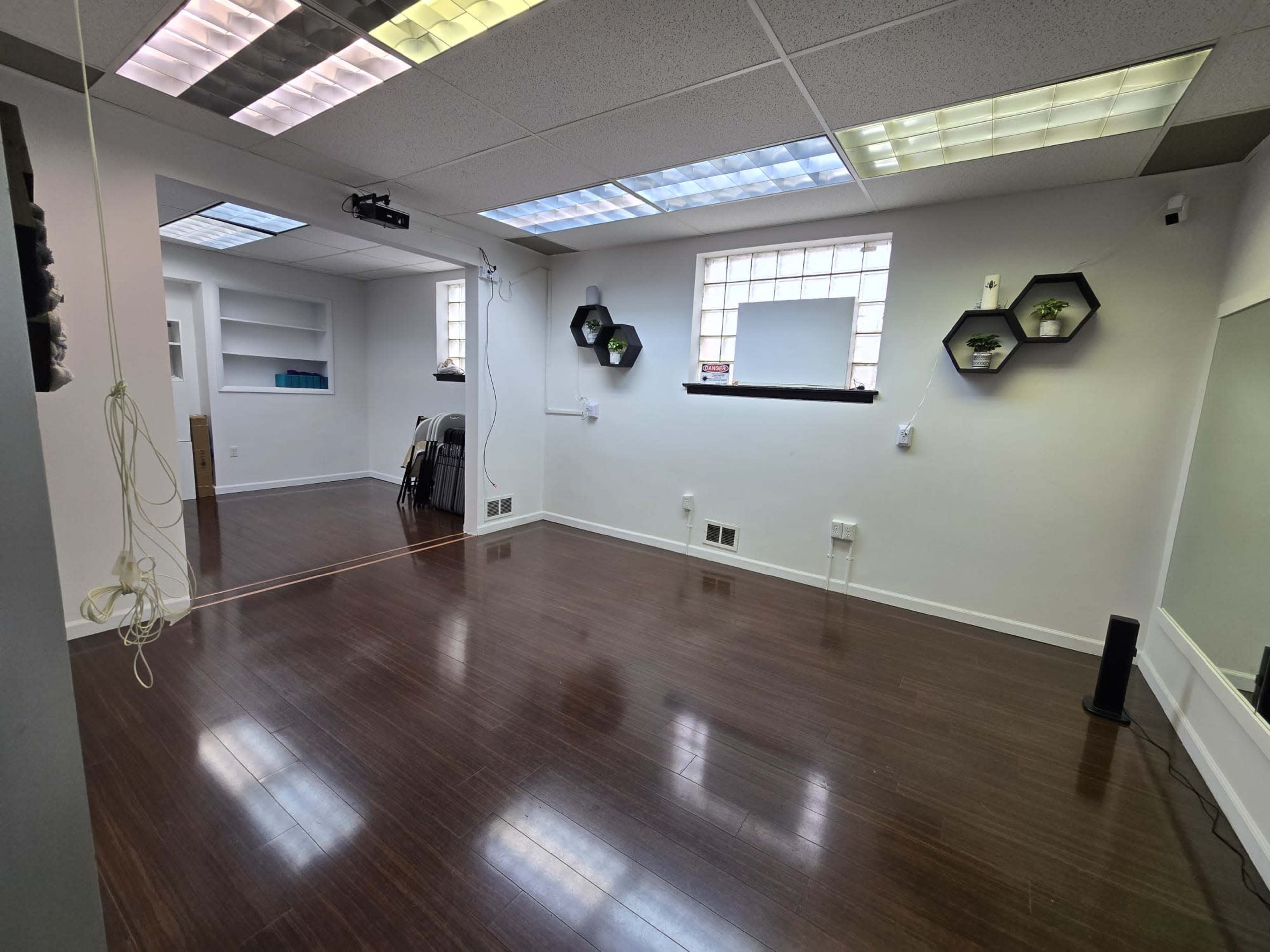 A bright, empty room with wooden flooring, two windows, and a few wall-mounted shelves.