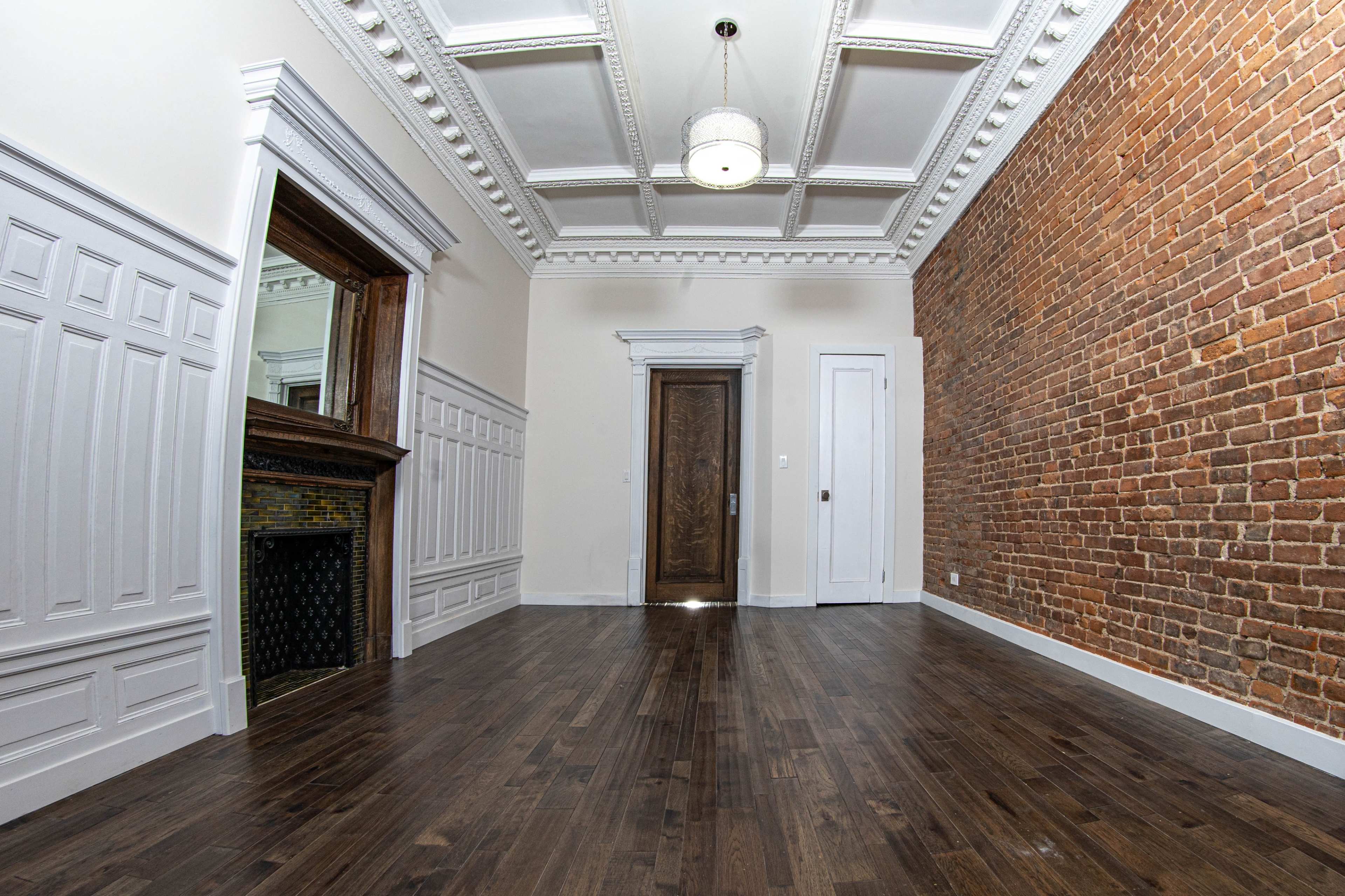 A spacious room featuring a mix of white paneling and a brick wall, with a wooden floor and a decorative ceiling.