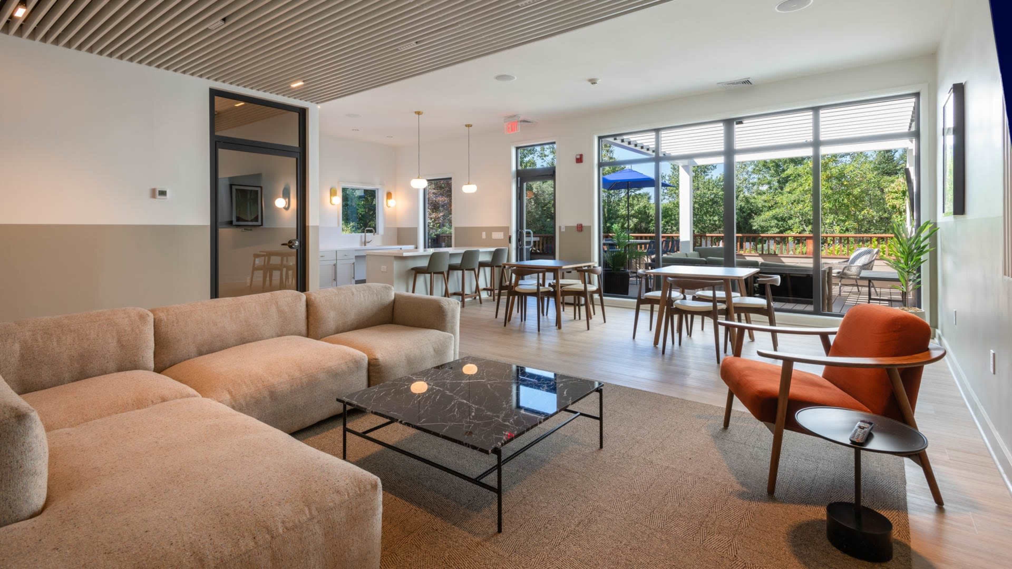 The image shows a modern lounge area with a spacious seating arrangement, a coffee table, and dining tables set near large windows overlooking greenery.