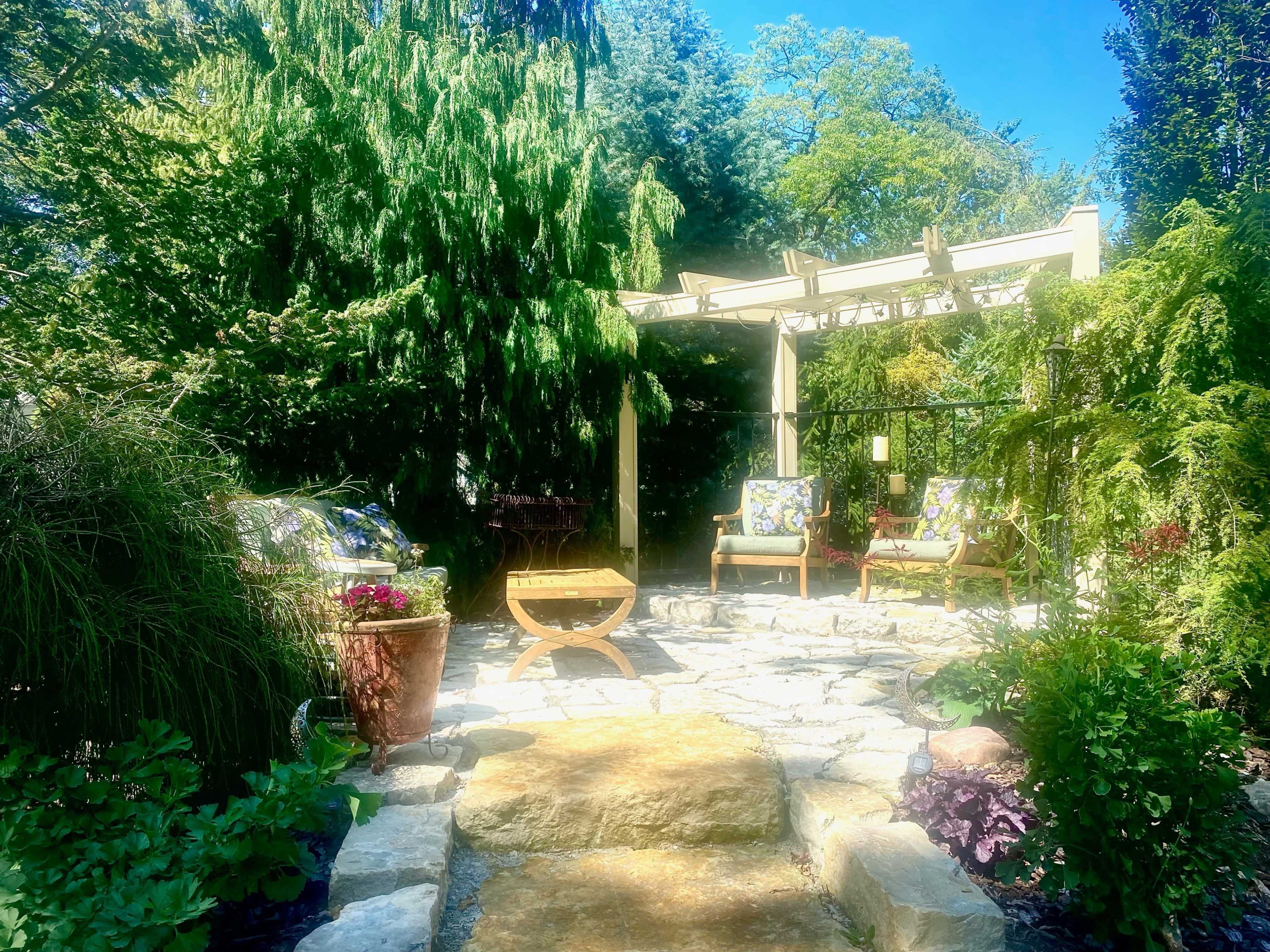 A stone pathway leads to a pergola surrounded by lush greenery and two chairs with floral cushions.