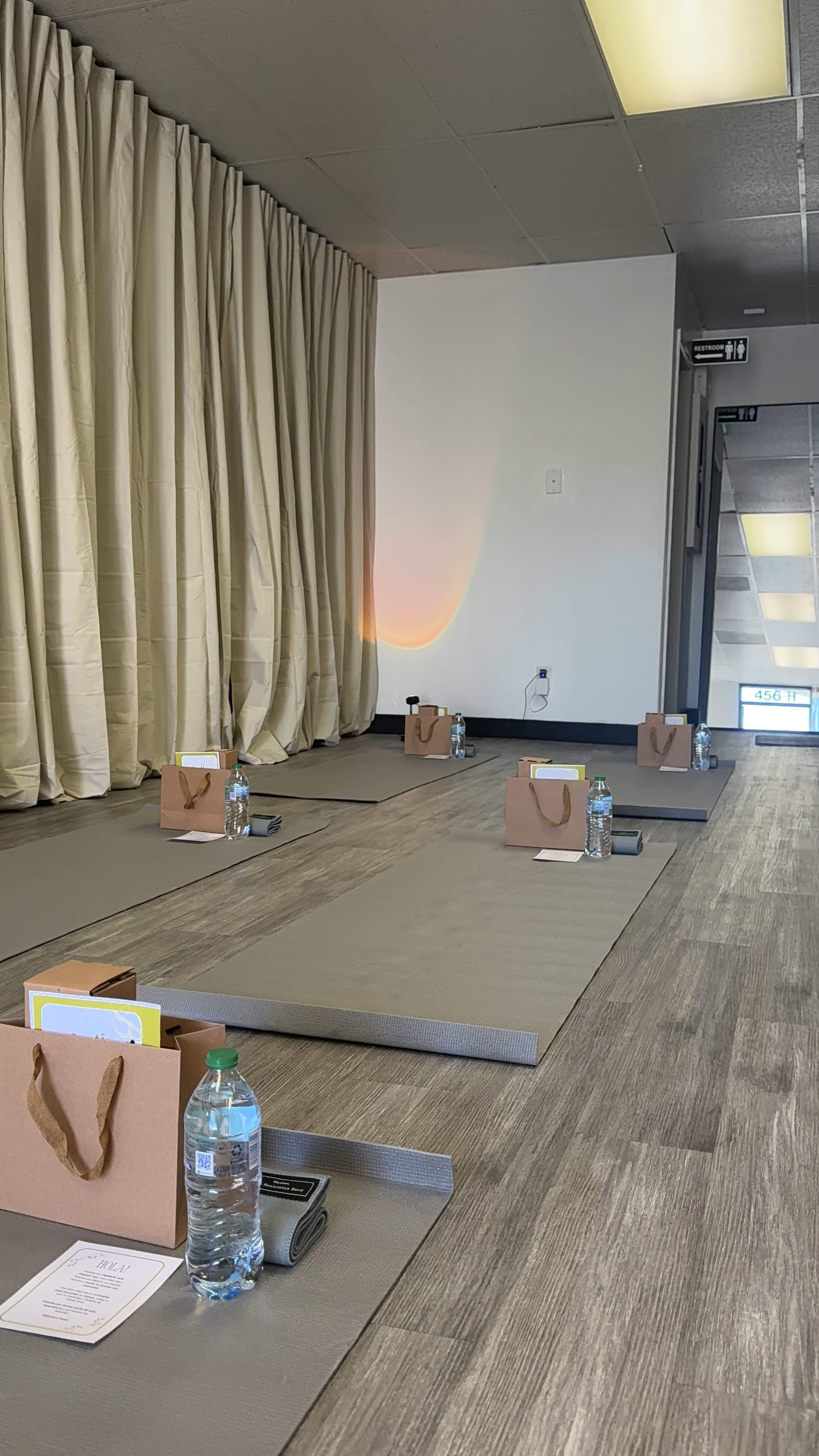 The image shows a yoga or meditation studio with neatly arranged mats, each accompanied by a brown bag and a water bottle, positioned in front of a curtain.