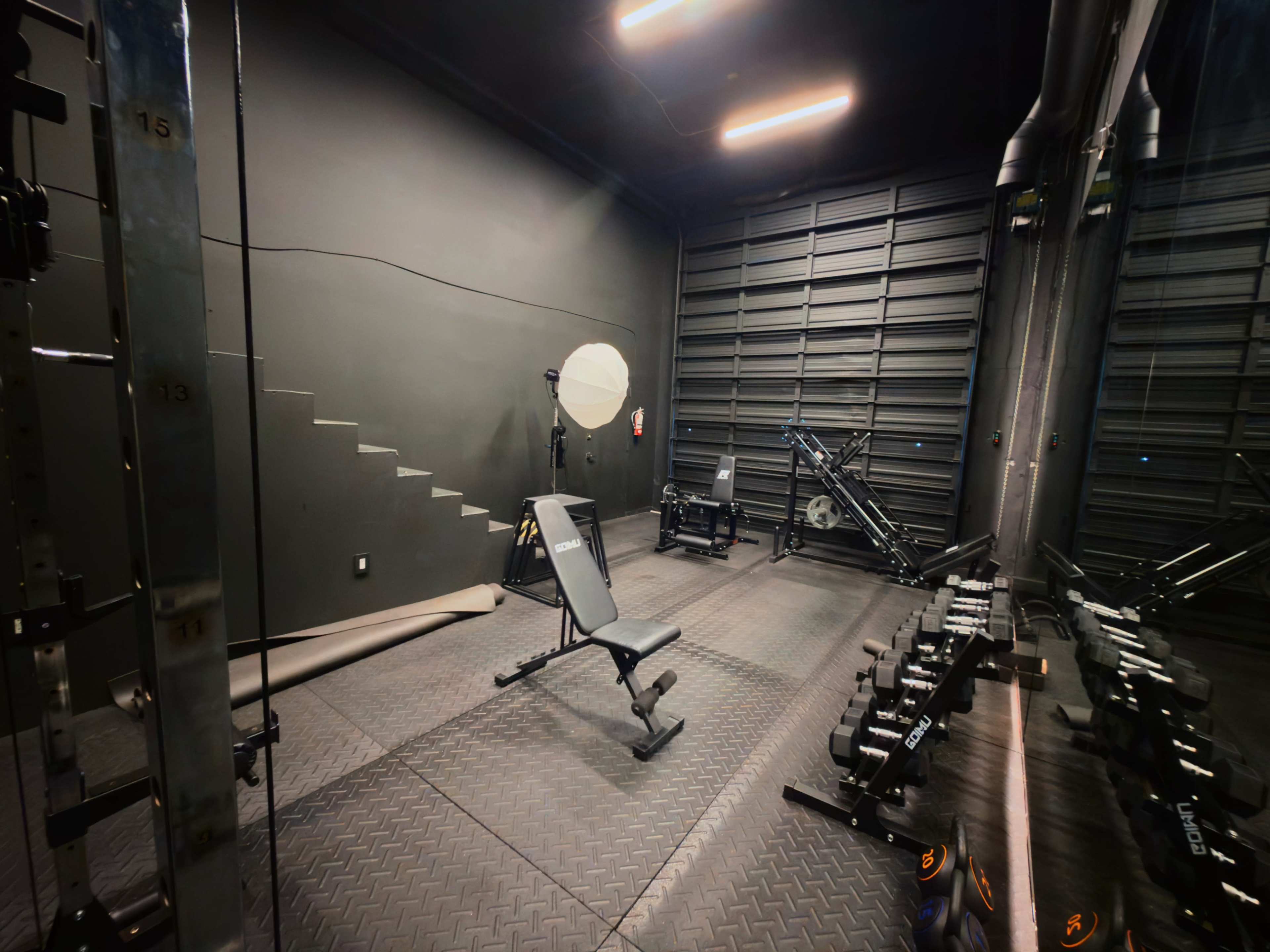 The image shows a small, dimly lit gym with various exercise equipment, including a weight bench, dumbbells, and a cable machine, set against a dark backdrop.