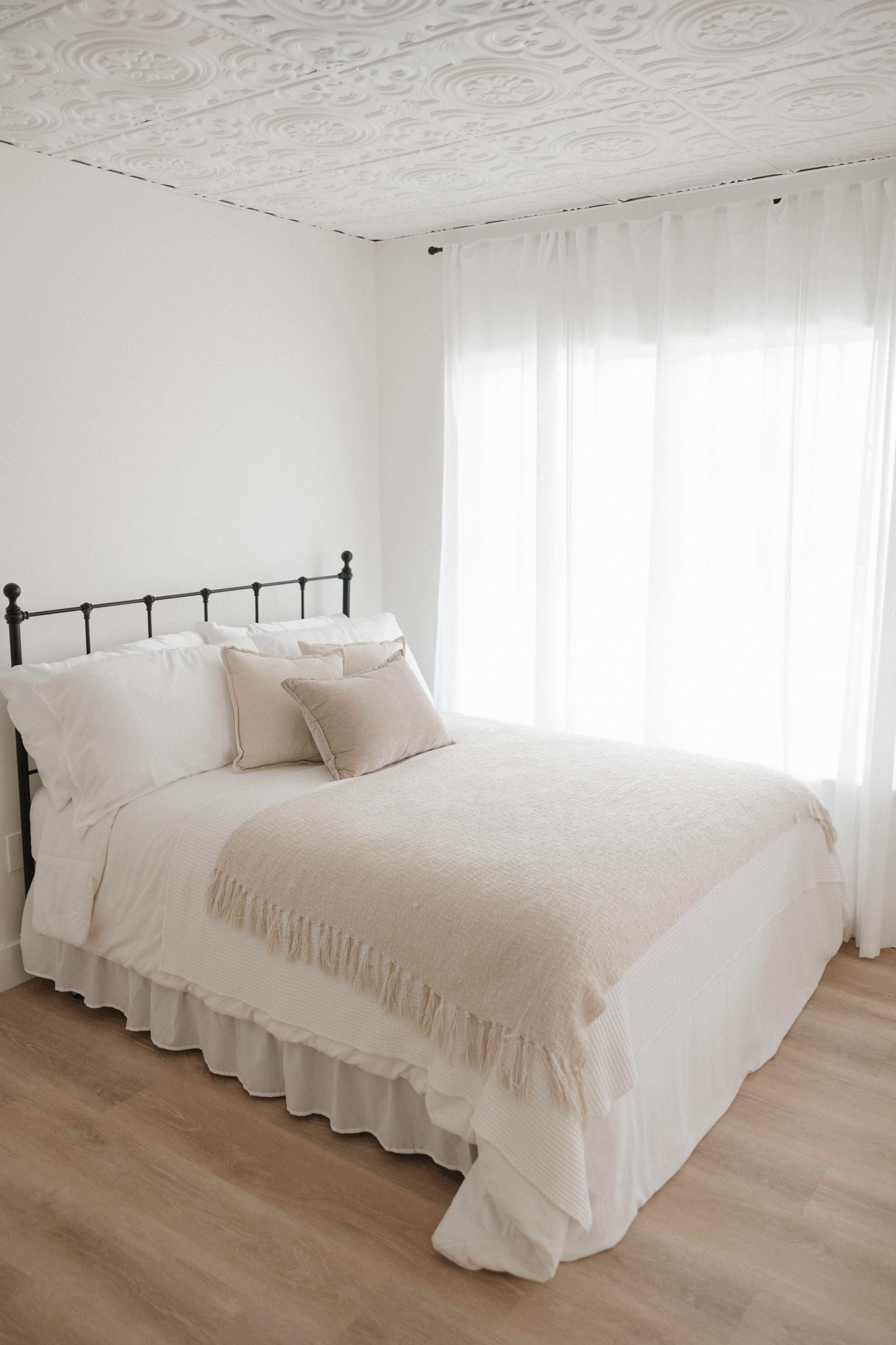 The image shows a minimalist bedroom with a black iron bed covered in white bedding and a tan throw blanket, positioned near a window with sheer curtains.