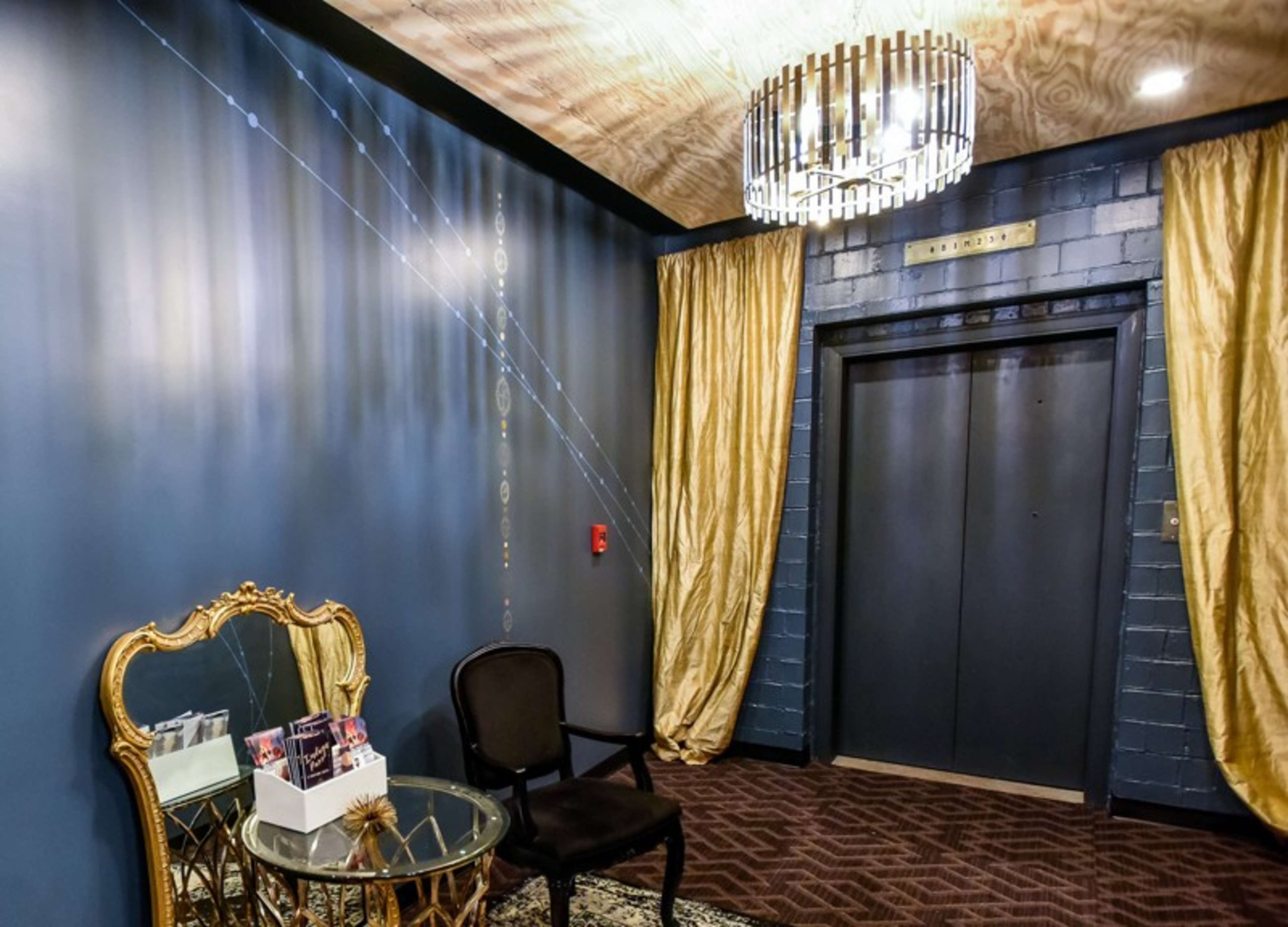 The image shows a hallway with a decorative chandelier, an elevator door, gold curtains, and a small round table with a display of materials, accompanied by a black chair.