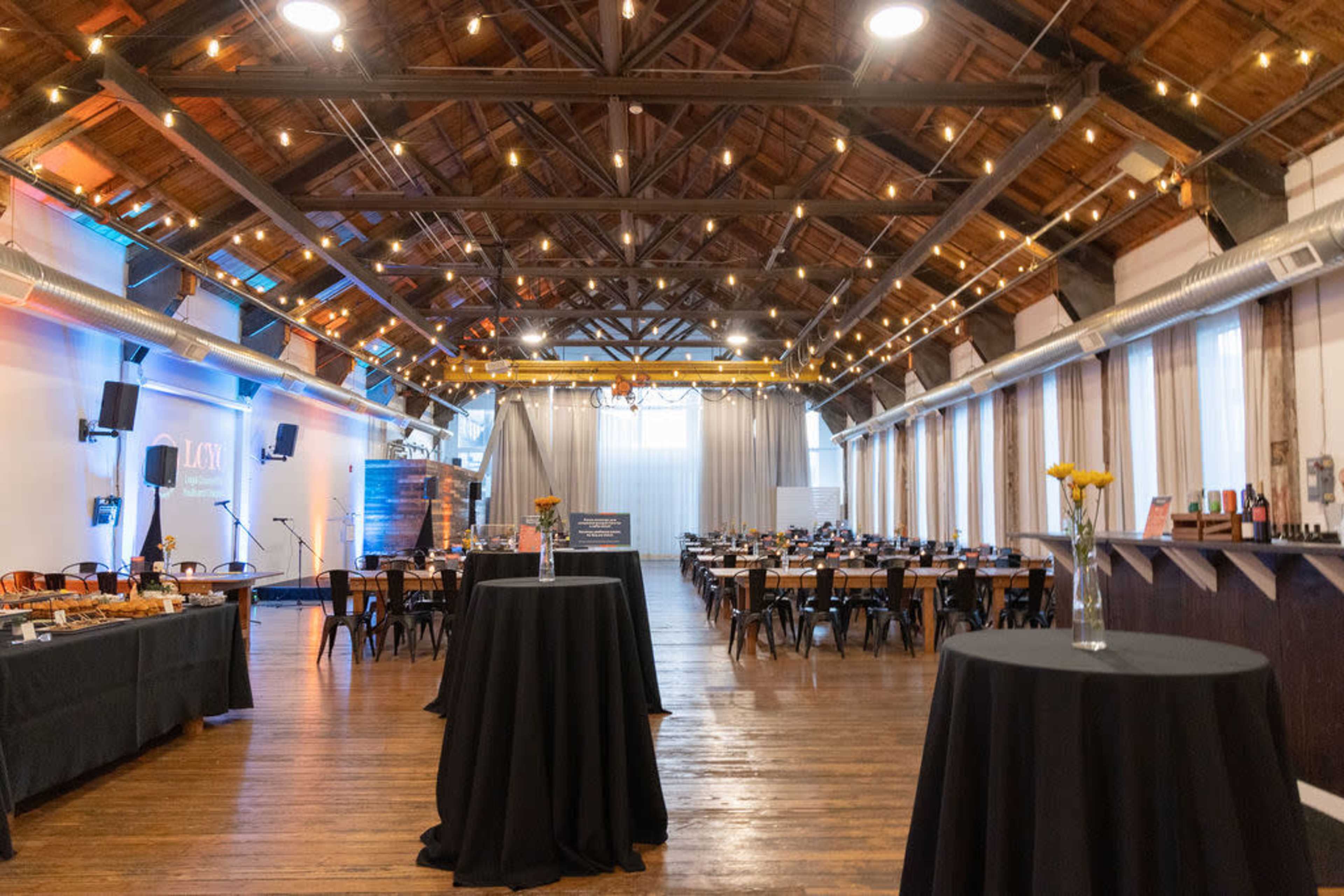 The image shows a spacious event venue with wooden floors, a high ceiling, and tables set for a gathering, illuminated by string lights.