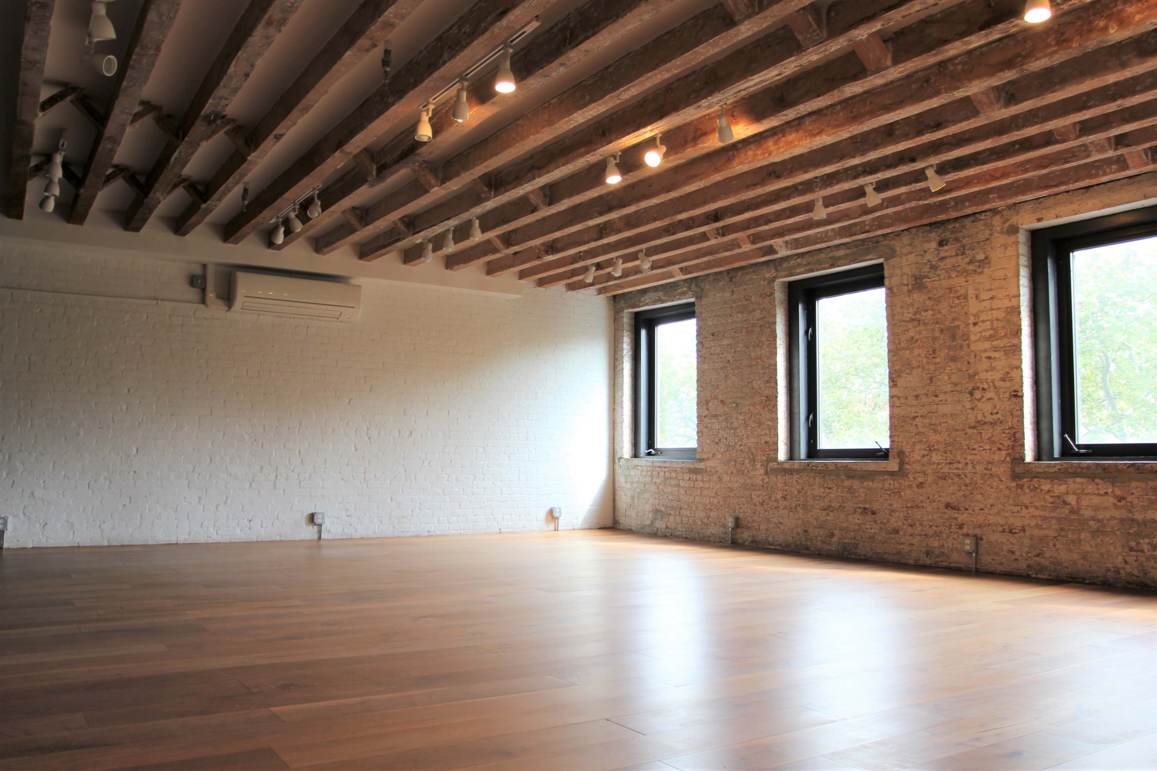 An empty room with wooden floorboards, exposed brick walls, and ceiling beams, featuring three windows that let in natural light.